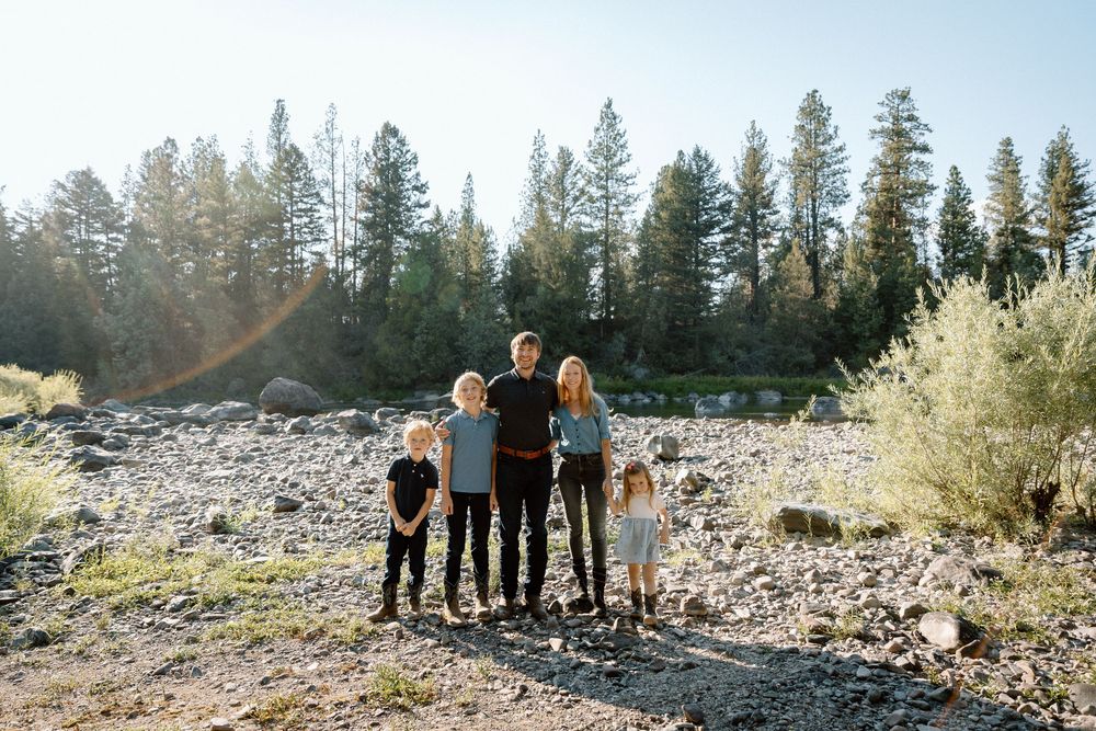 Family of five posing outdoors on rocky terrain surrounded by tall pine trees with warm sunlight in background.