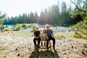 Three children sit together on rocks in a sunny forest clearing with tall pine trees and bright sunlight in the background.