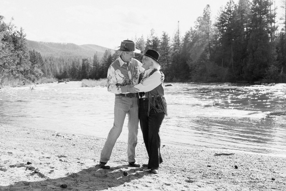 Two people embracing on a sandy riverbank with mountains and pine trees in a black and white photo.