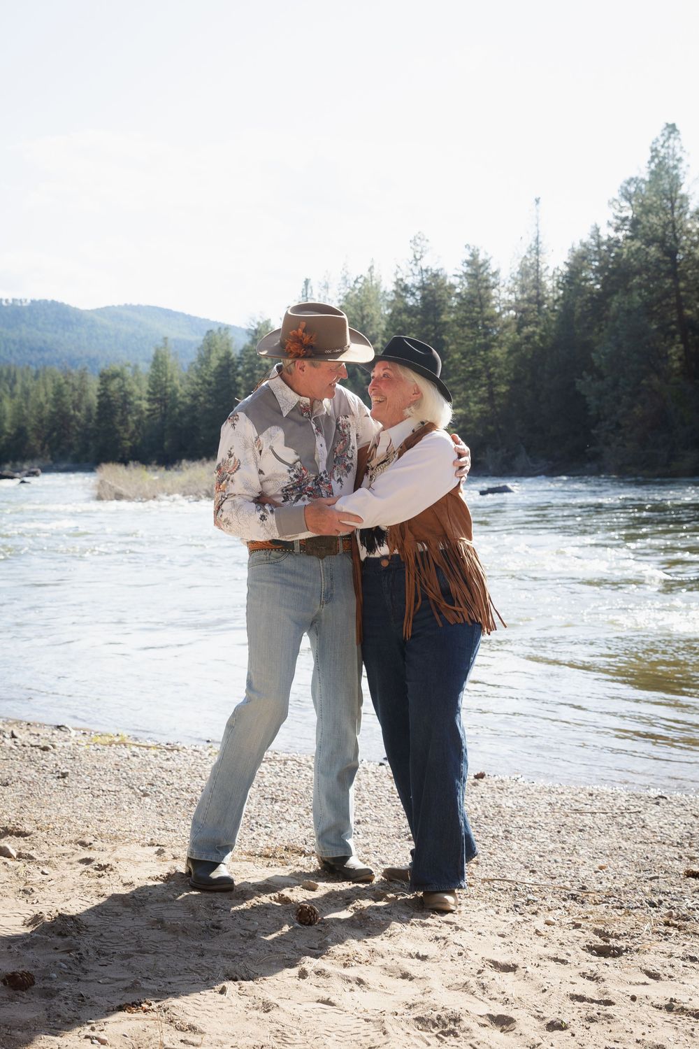 Elderly couple in cowboy hats dancing together on a riverbank surrounded by pine trees and mountains.