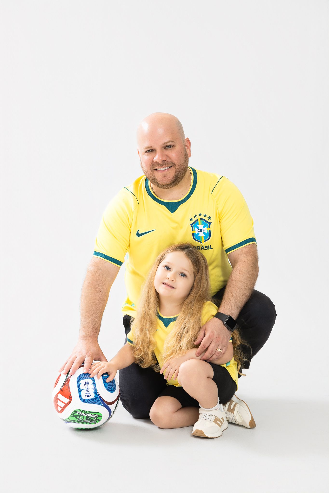 A bald man and young girl in matching Brazil soccer jerseys pose with a soccer ball on a white background.