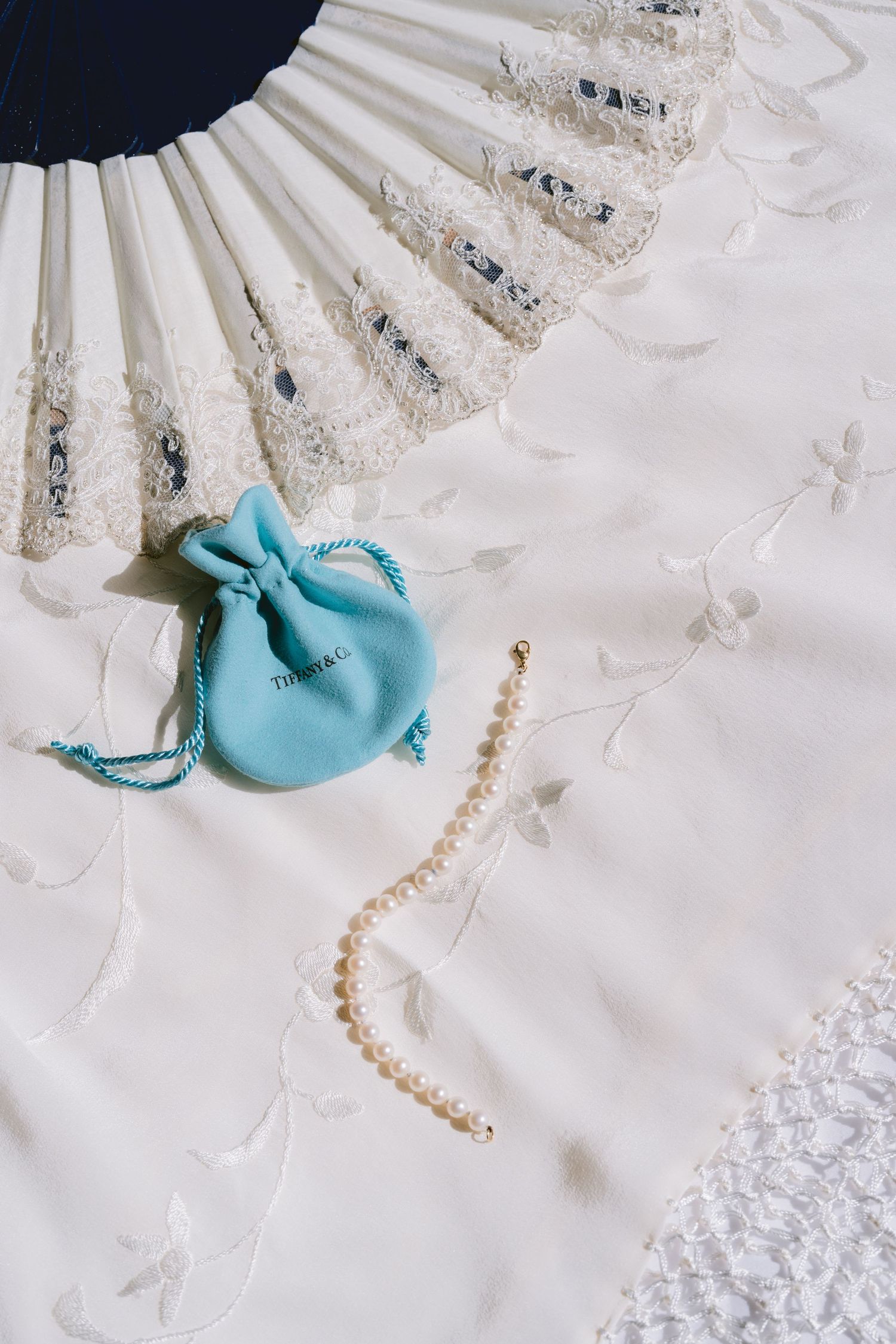Tiffany & Co. blue pouch with pearl bracelet on white lace wedding dress fabric with decorative fan.