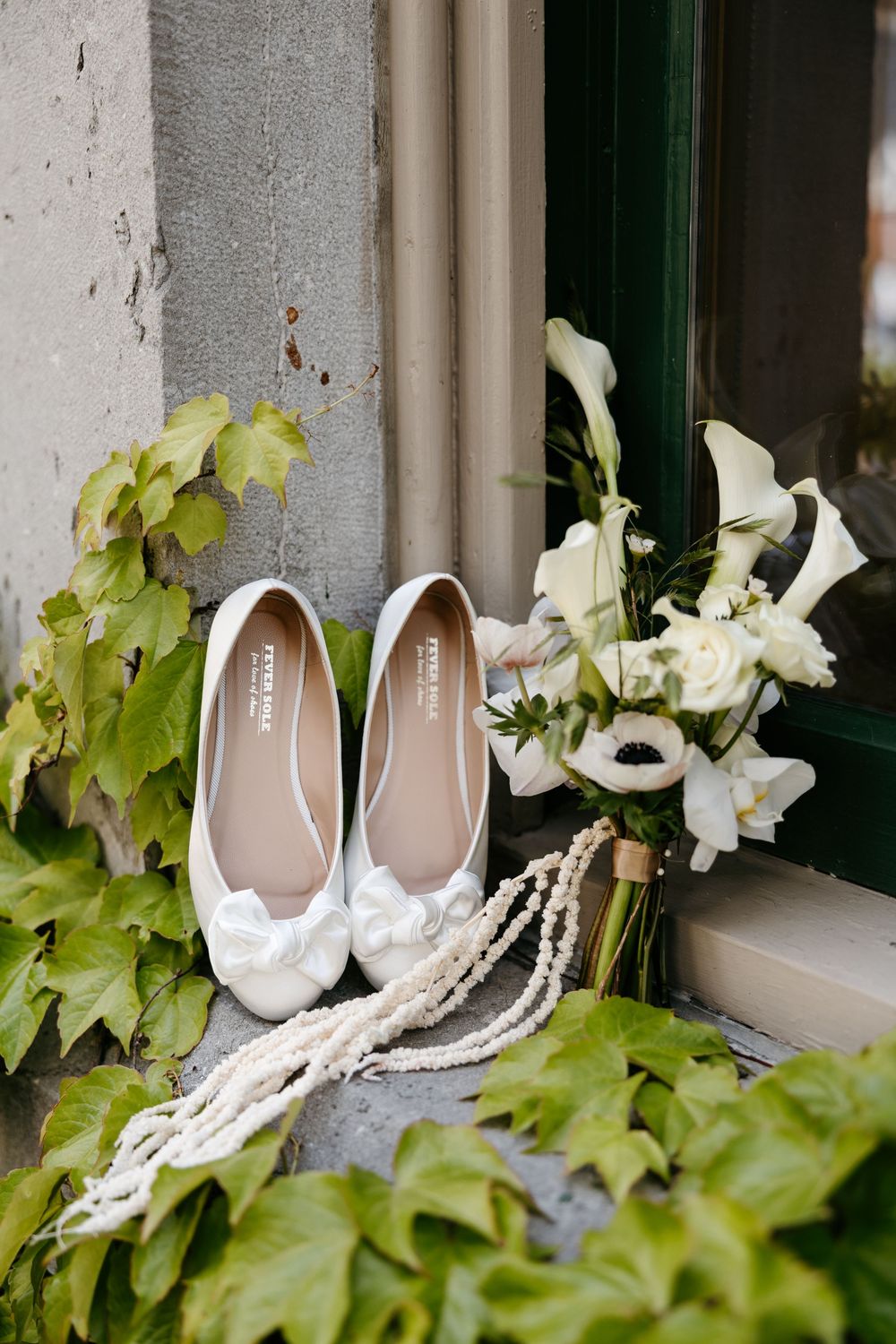 White bridal ballet flats with bow detail beside pearl necklace and white calla lily bouquet on stone steps.