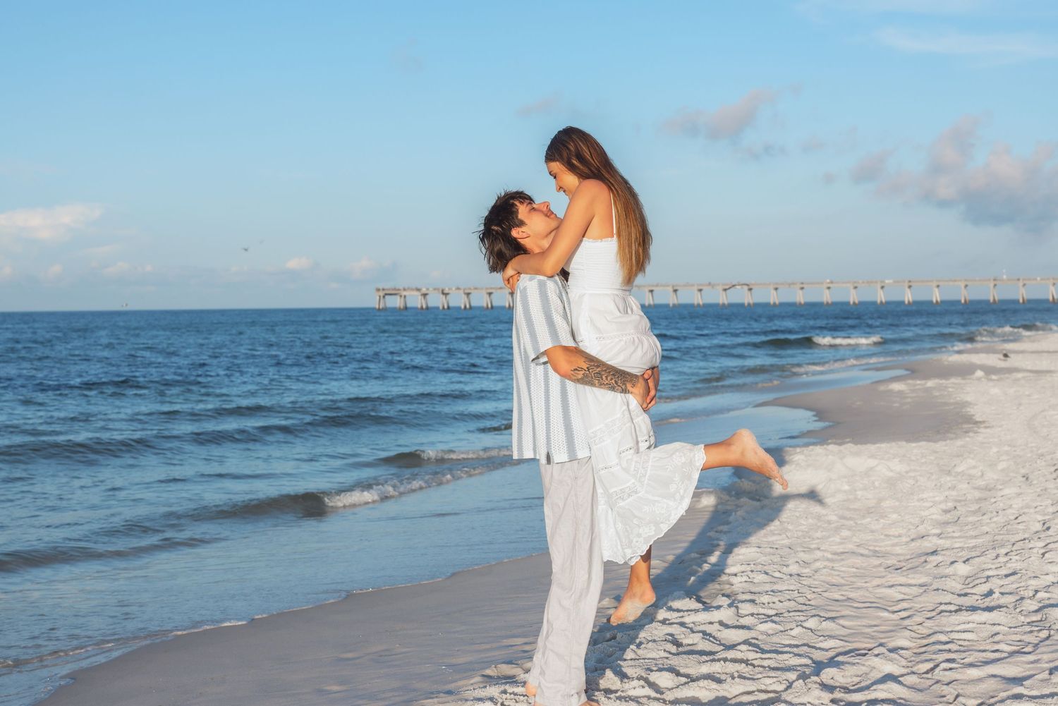 Romantic couple embracing on a white sand beach, man lifting woman in white dress near ocean waves.