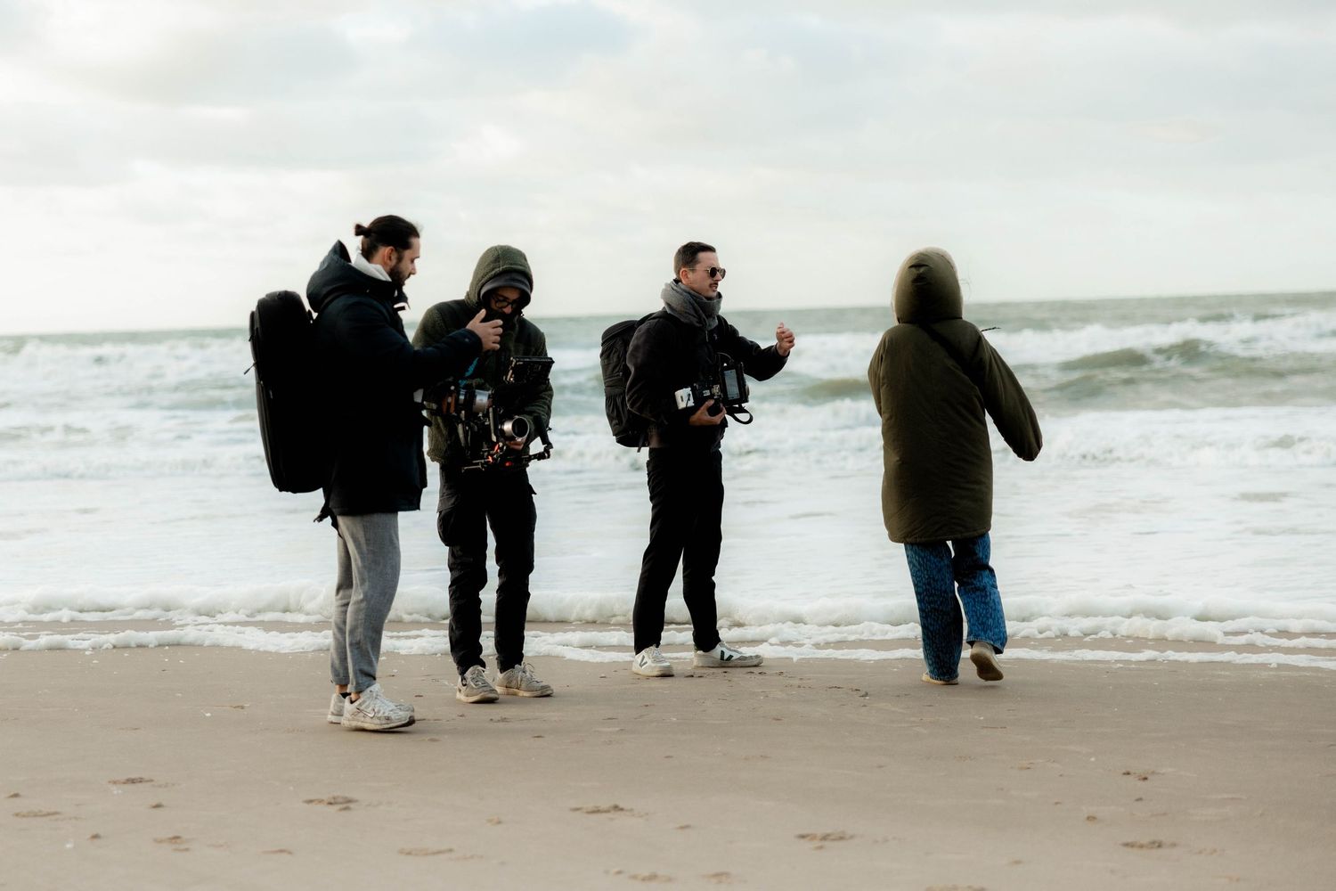 Two men with a video camera filming on a windy beach with ocean waves in the background.