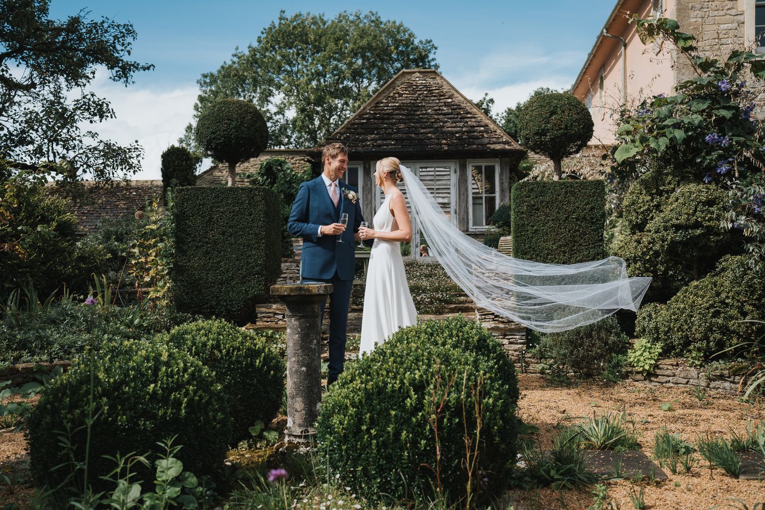 Bride and groom stand in a formal garden with topiary hedges, brides veil flowing in the wind on a sunny day.