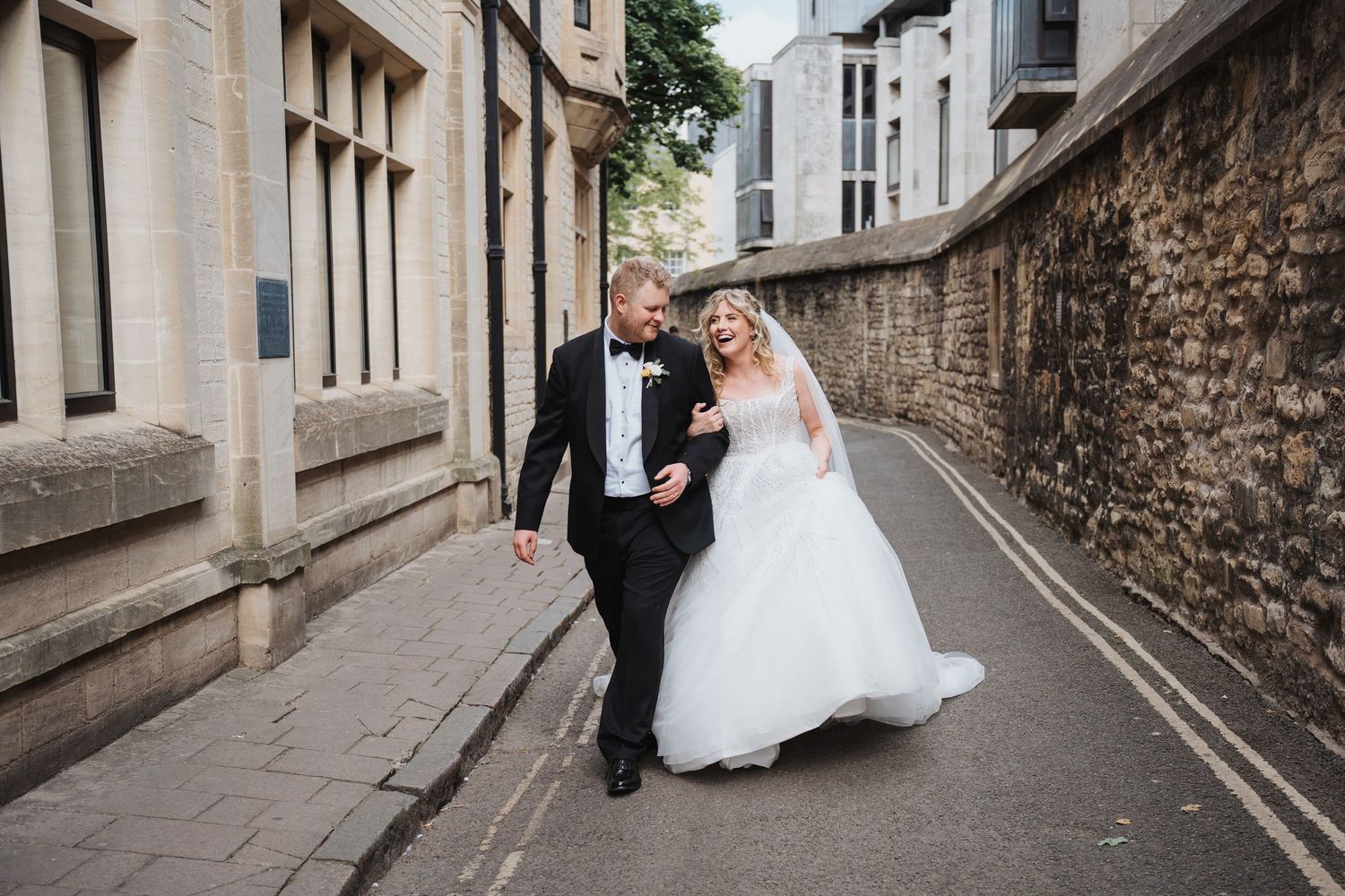 Bride and groom walking and laughing together on a cobblestone street lined with stone buildings on their wedding day.
