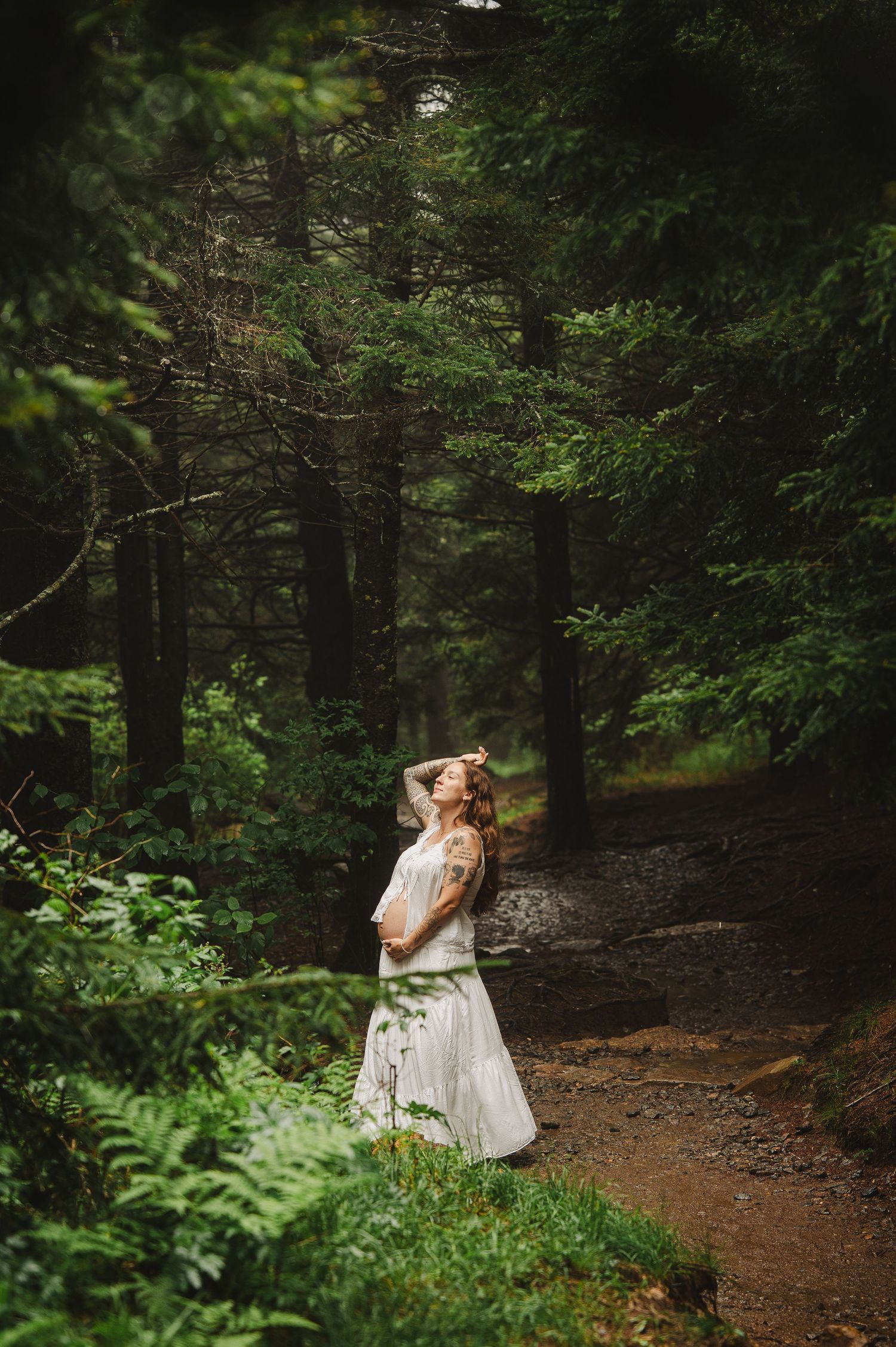Bride in white gown standing on a forest path, looking up through tall green trees in a serene woodland setting.
