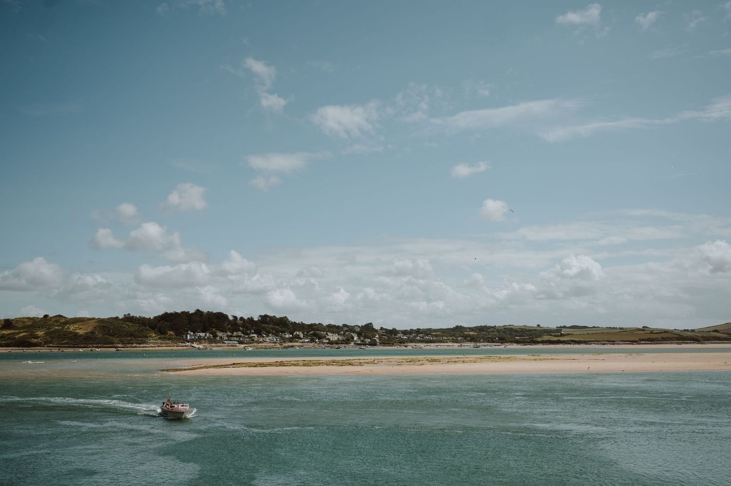 A small boat cruises along a calm turquoise bay with a sandy beach and green coastal village under a partly cloudy sky.