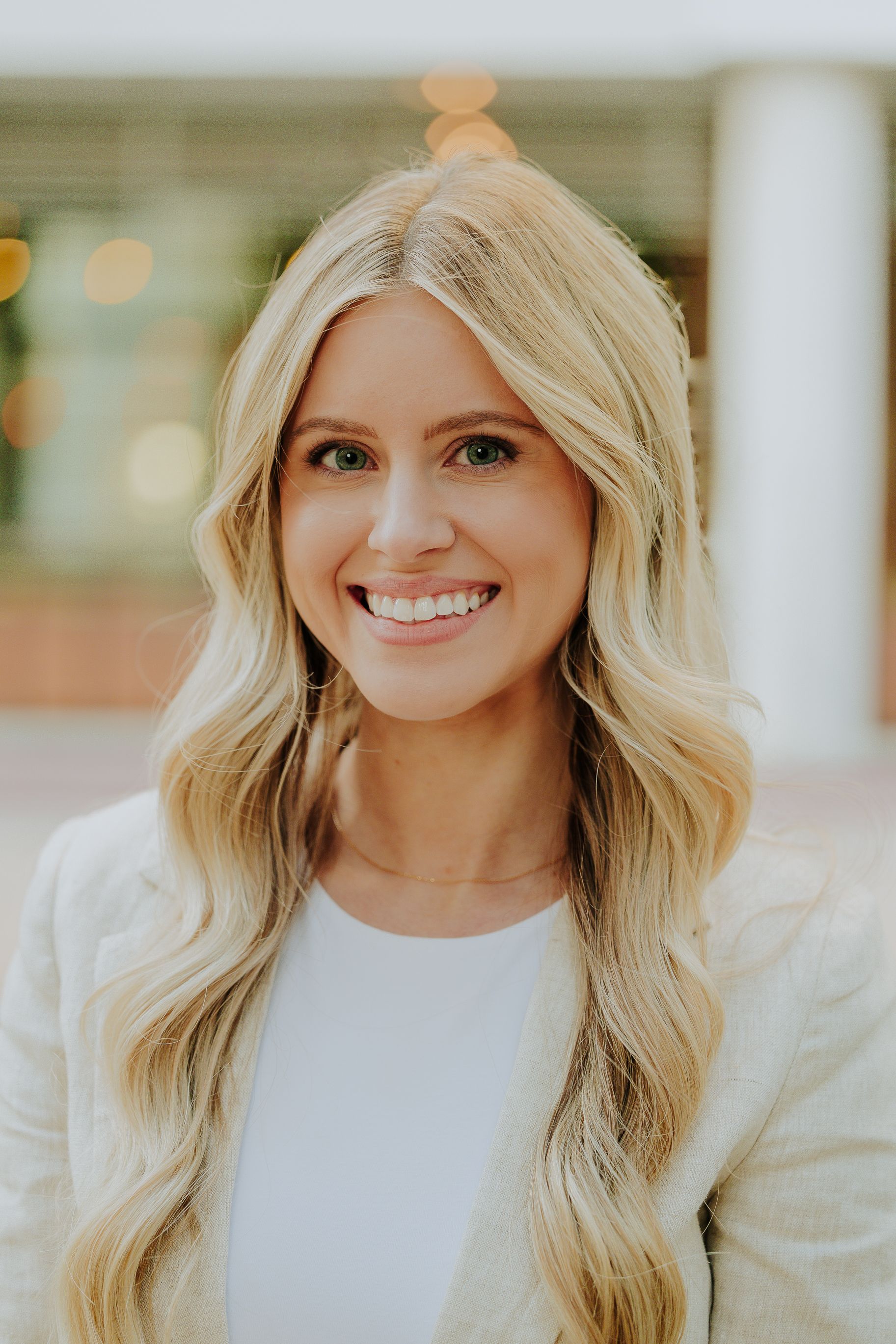 Smiling blonde woman in a beige blazer poses for a professional headshot outdoors.