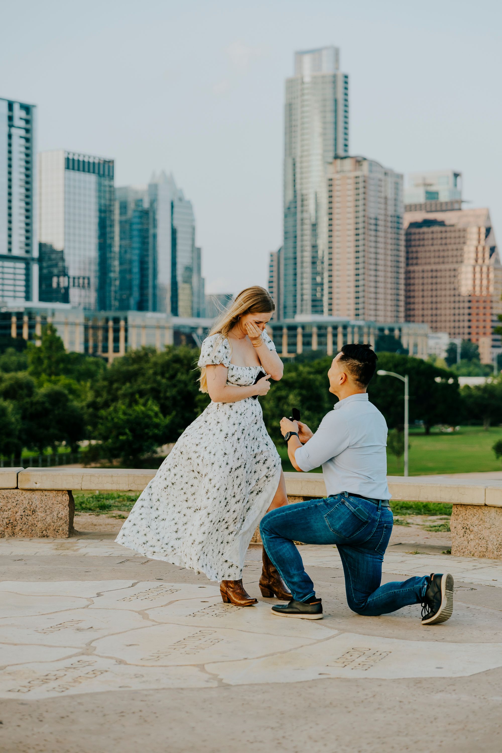 Man proposes on one knee to woman in floral dress with Austin Texas skyline in background.
