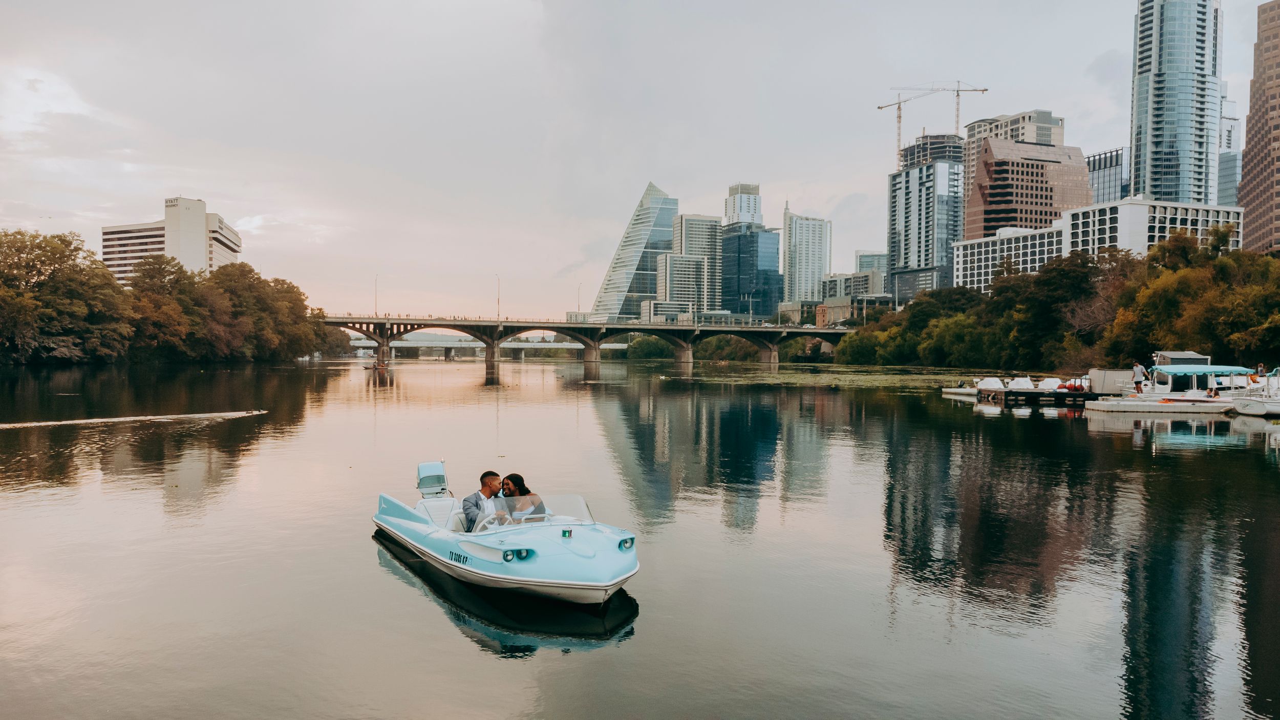 A couple enjoys a leisurely boat ride on a calm river with a city skyline and bridge in the background.