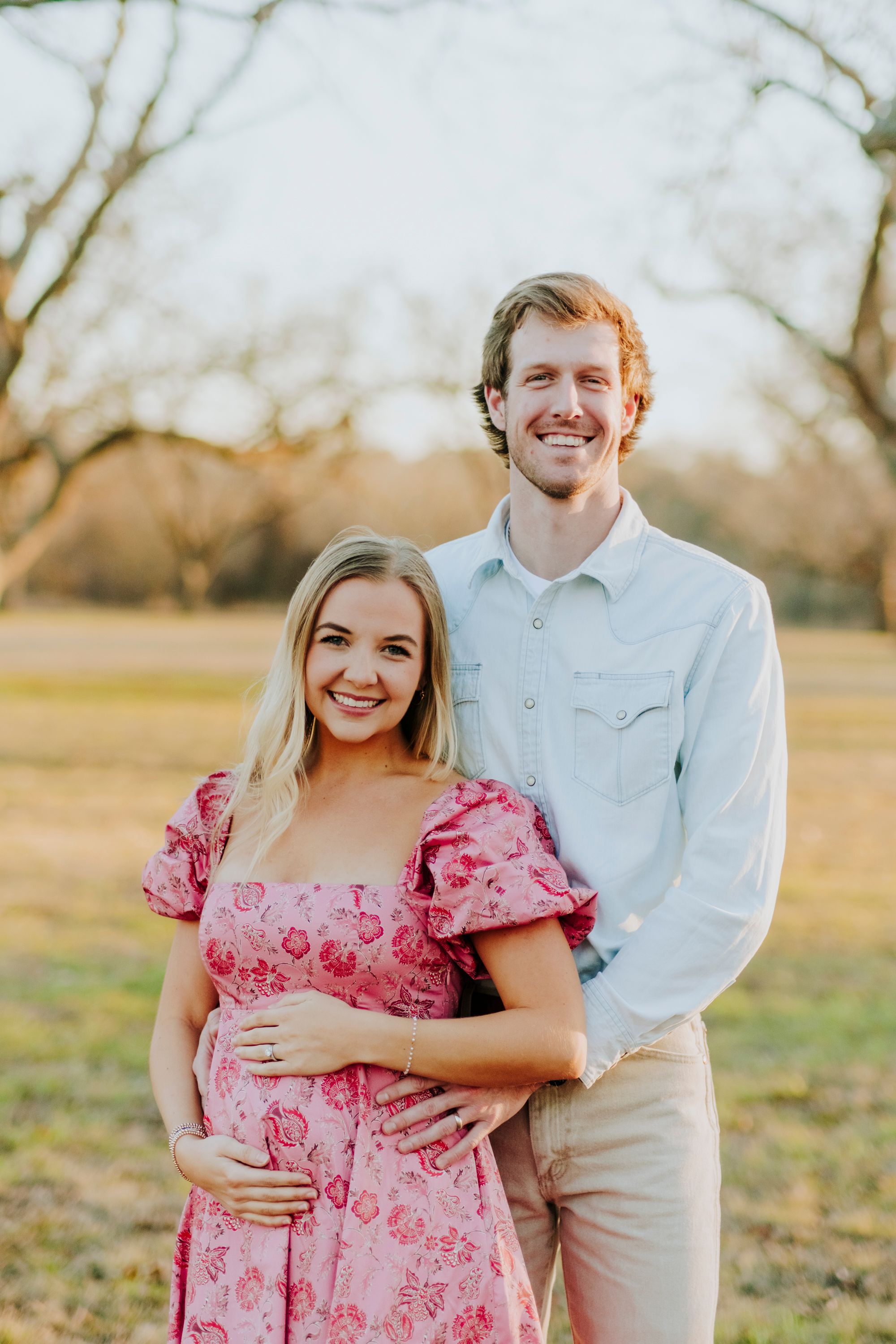 Happy couple embracing outdoors in a sunny field, woman wearing pink floral dress, man in light blue shirt smiling.