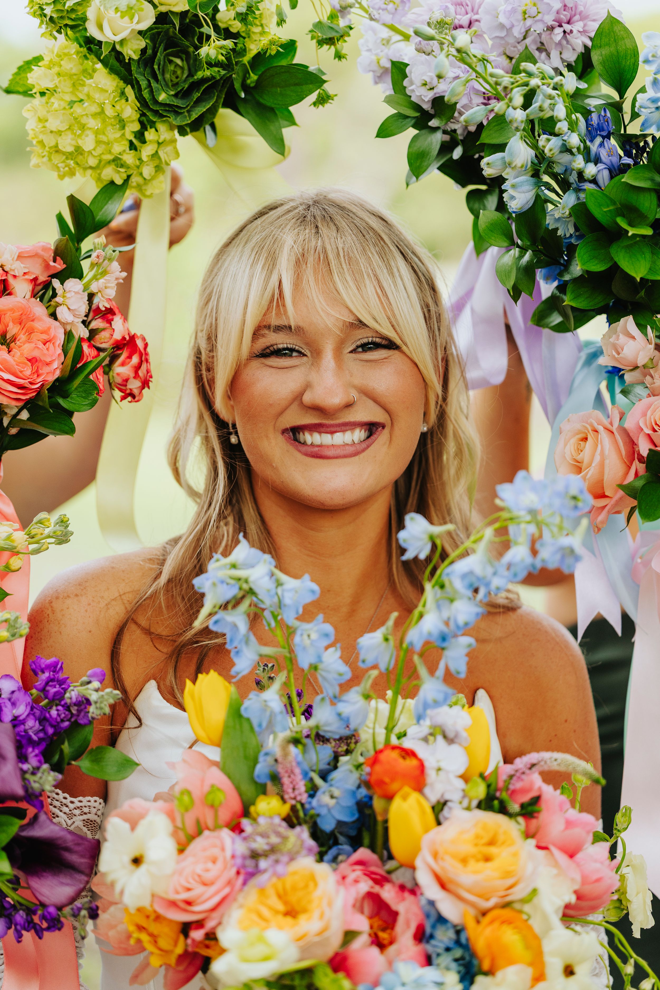Smiling bride with blonde bangs surrounded by colorful wedding bouquets featuring roses, tulips, and blue delphiniums.