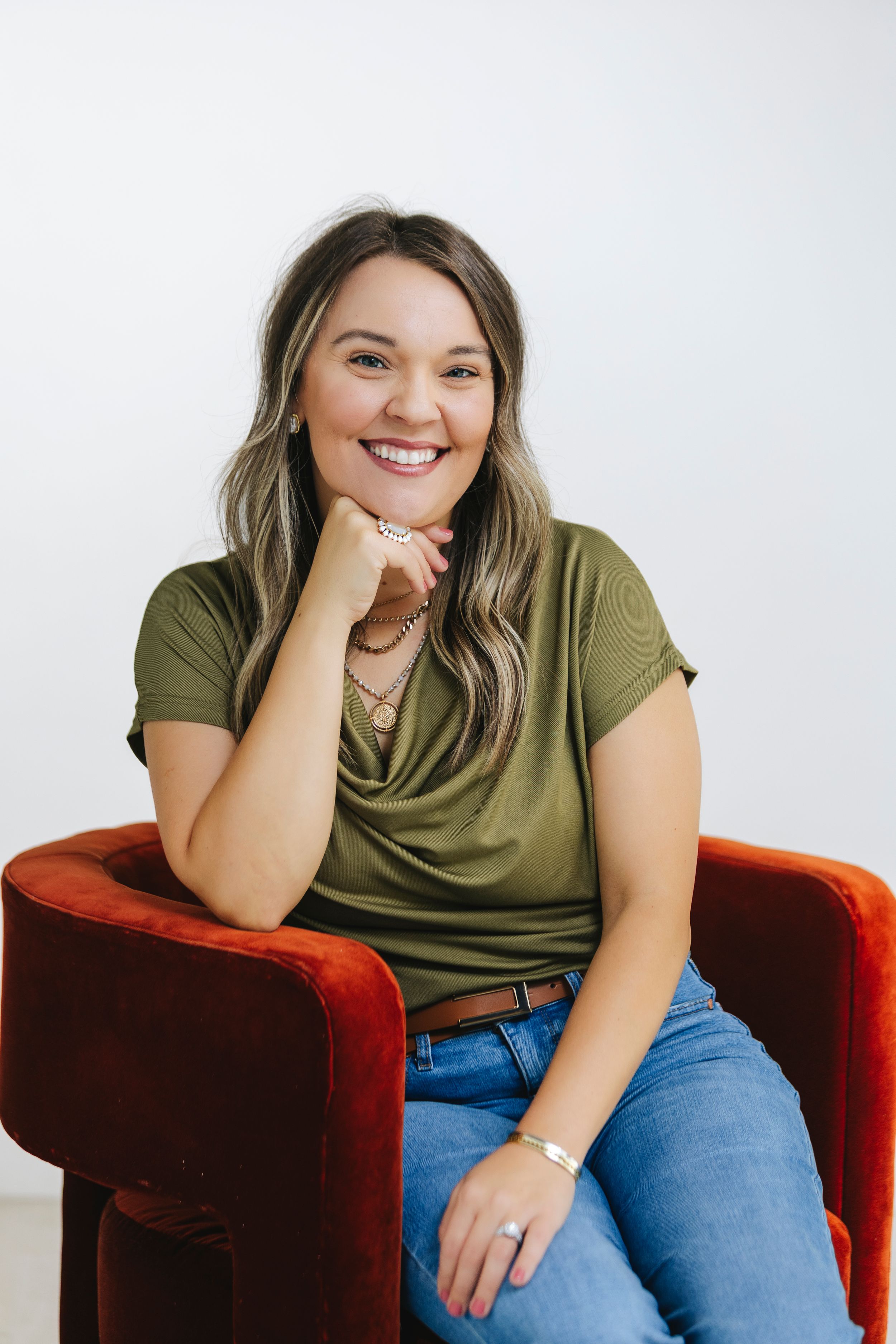 Smiling woman in green t-shirt and jeans sitting relaxed in a red velvet chair against a white background.