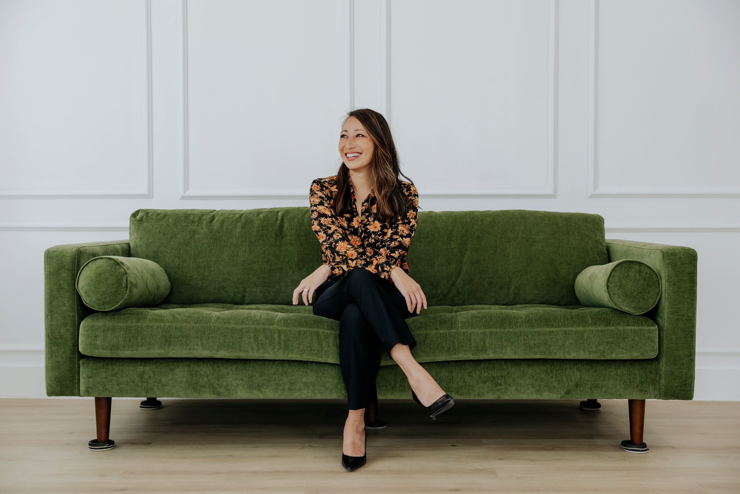 Woman in floral top sitting on green velvet sofa, smiling, in bright white room with hardwood floors.