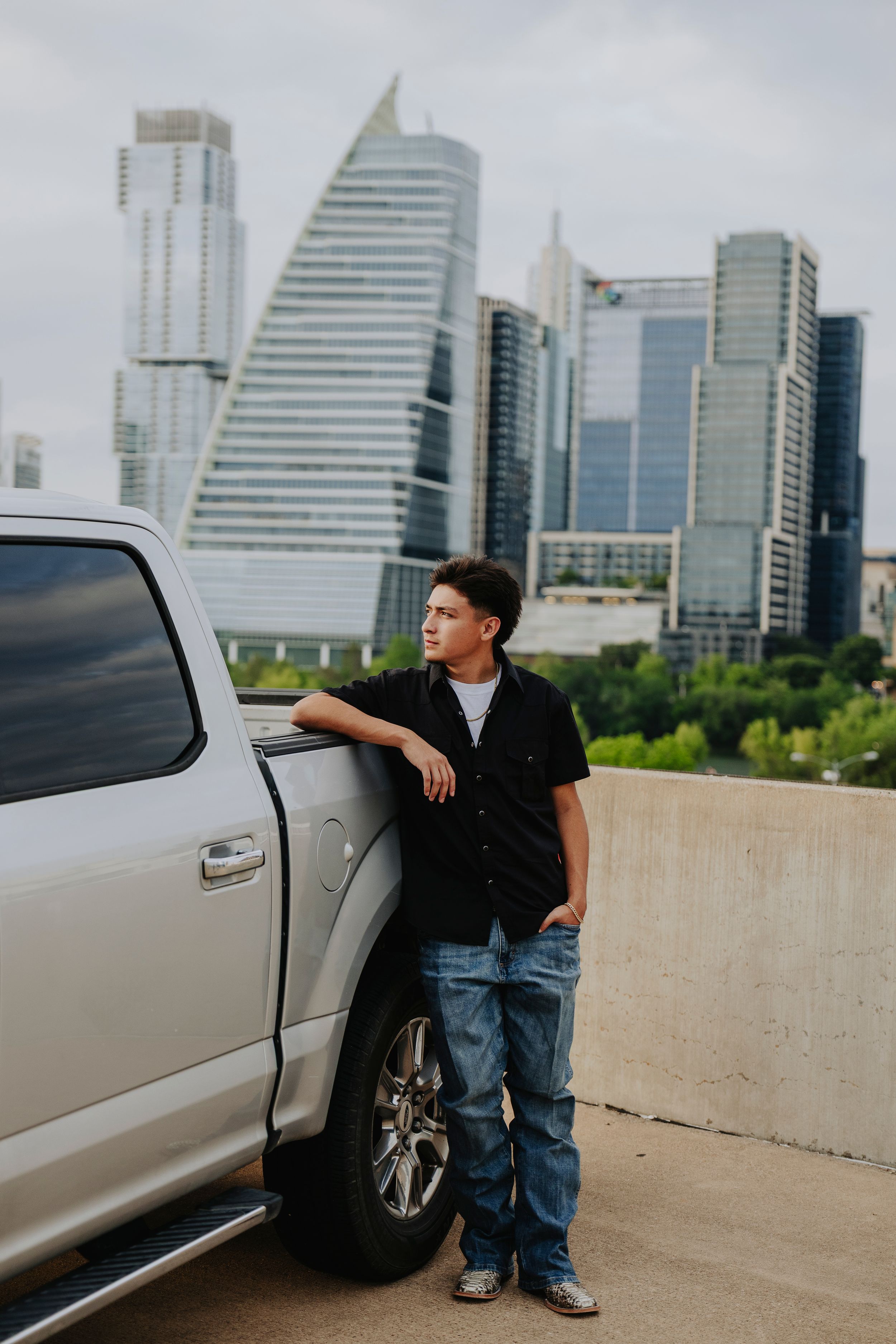 Young man in black shirt and jeans leaning on silver truck with city skyline in background.