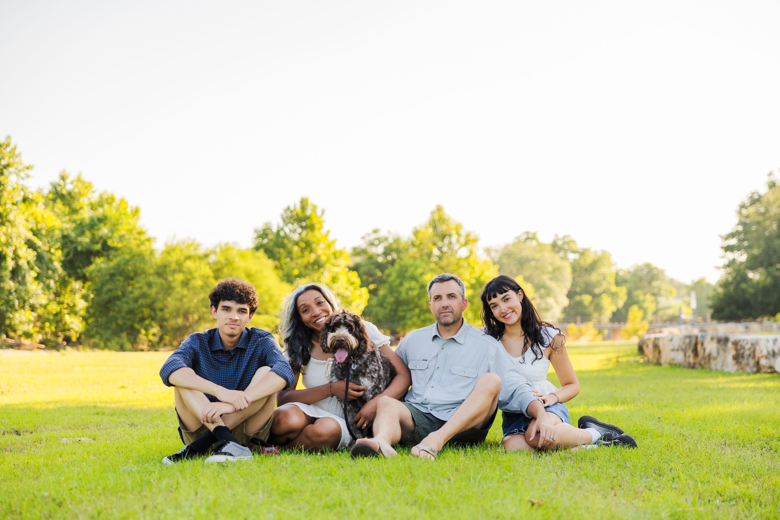 Family of four sitting on green grass with their dog in a sunny park surrounded by trees.