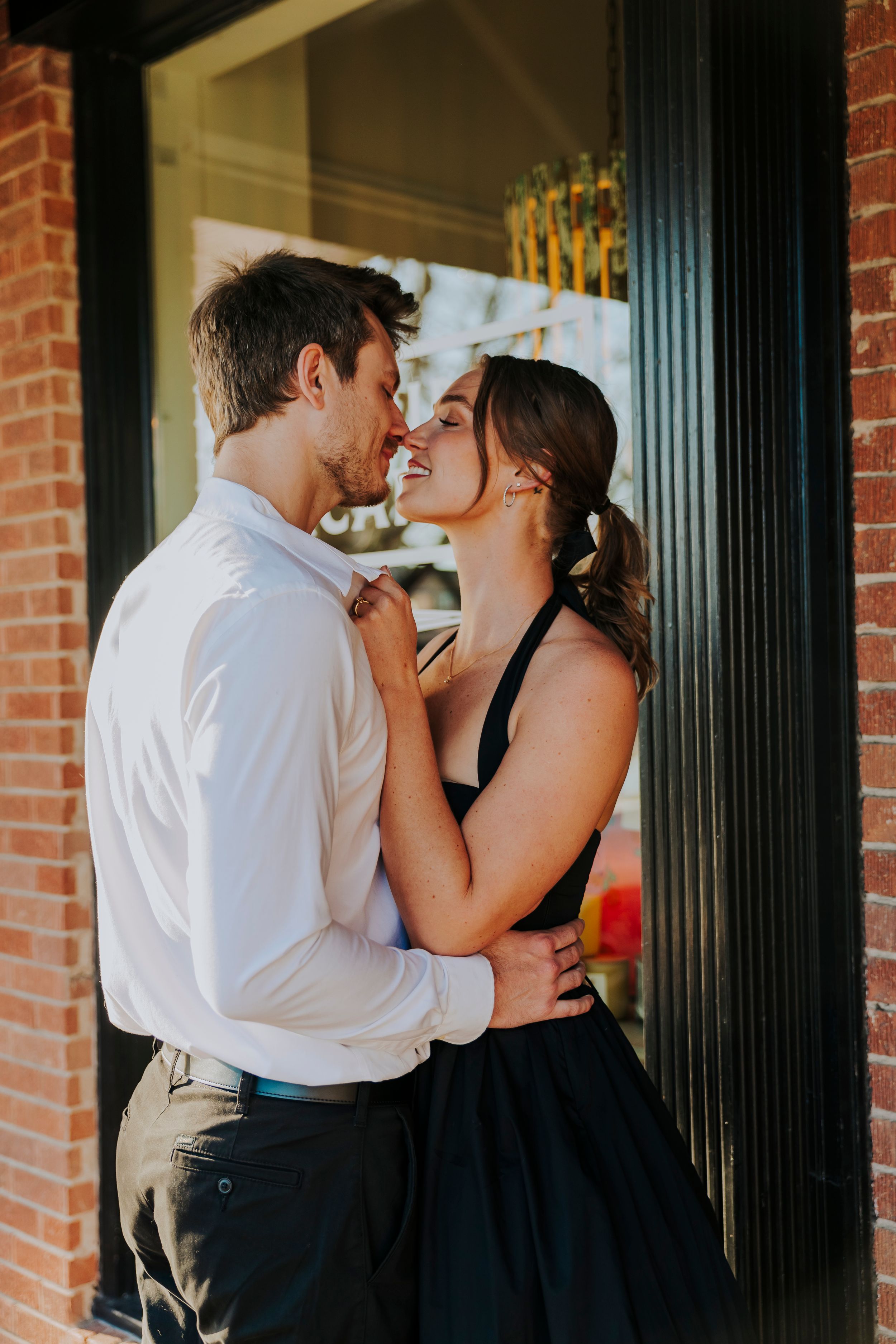Romantic couple embracing and almost kissing outdoors near brick wall, woman in black dress holding man's collar.