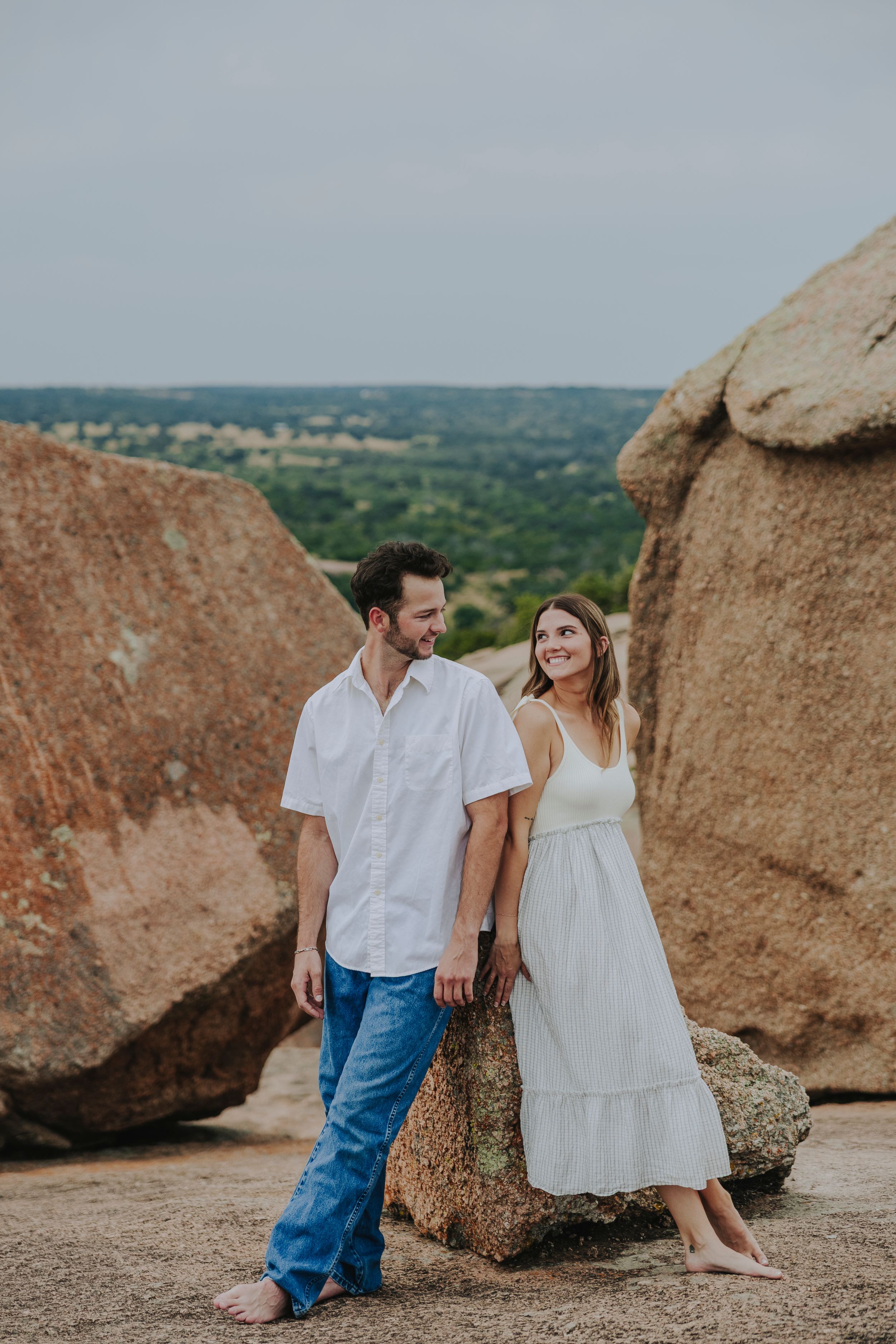 Couple in white outfits standing among large boulders at Enchanted Rock, smiling at each other with scenic Texas Hill Country views.