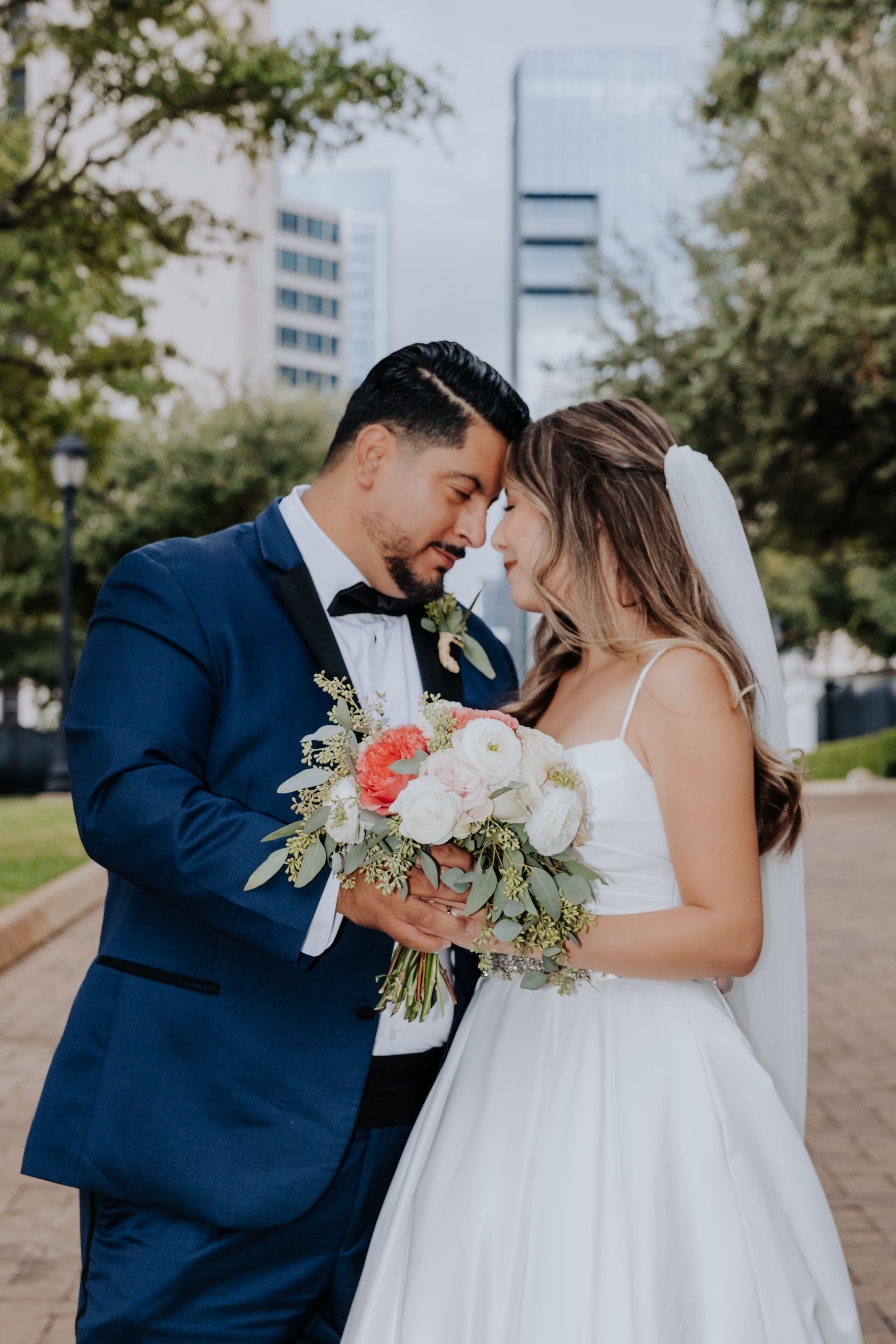 Bride and groom touching foreheads, holding white and coral bouquet, groom in navy suit, urban backdrop.