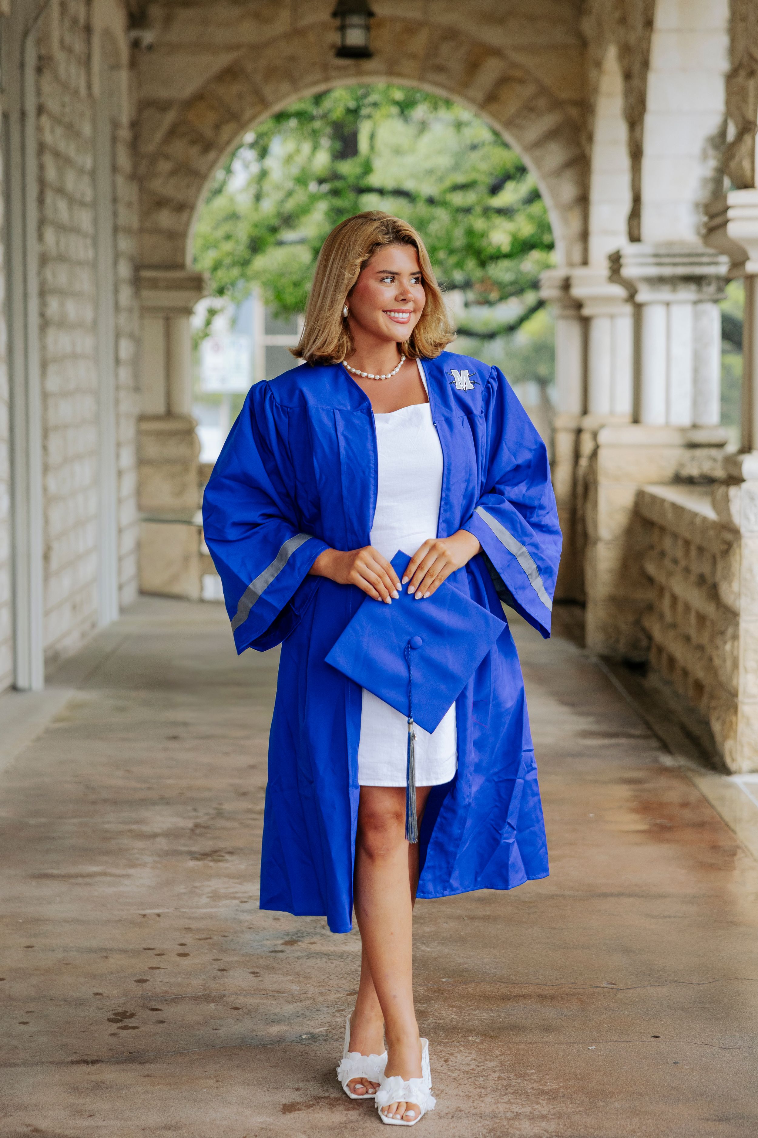 Young woman in royal blue graduation gown and cap smiling in an elegant stone archway colonnade.