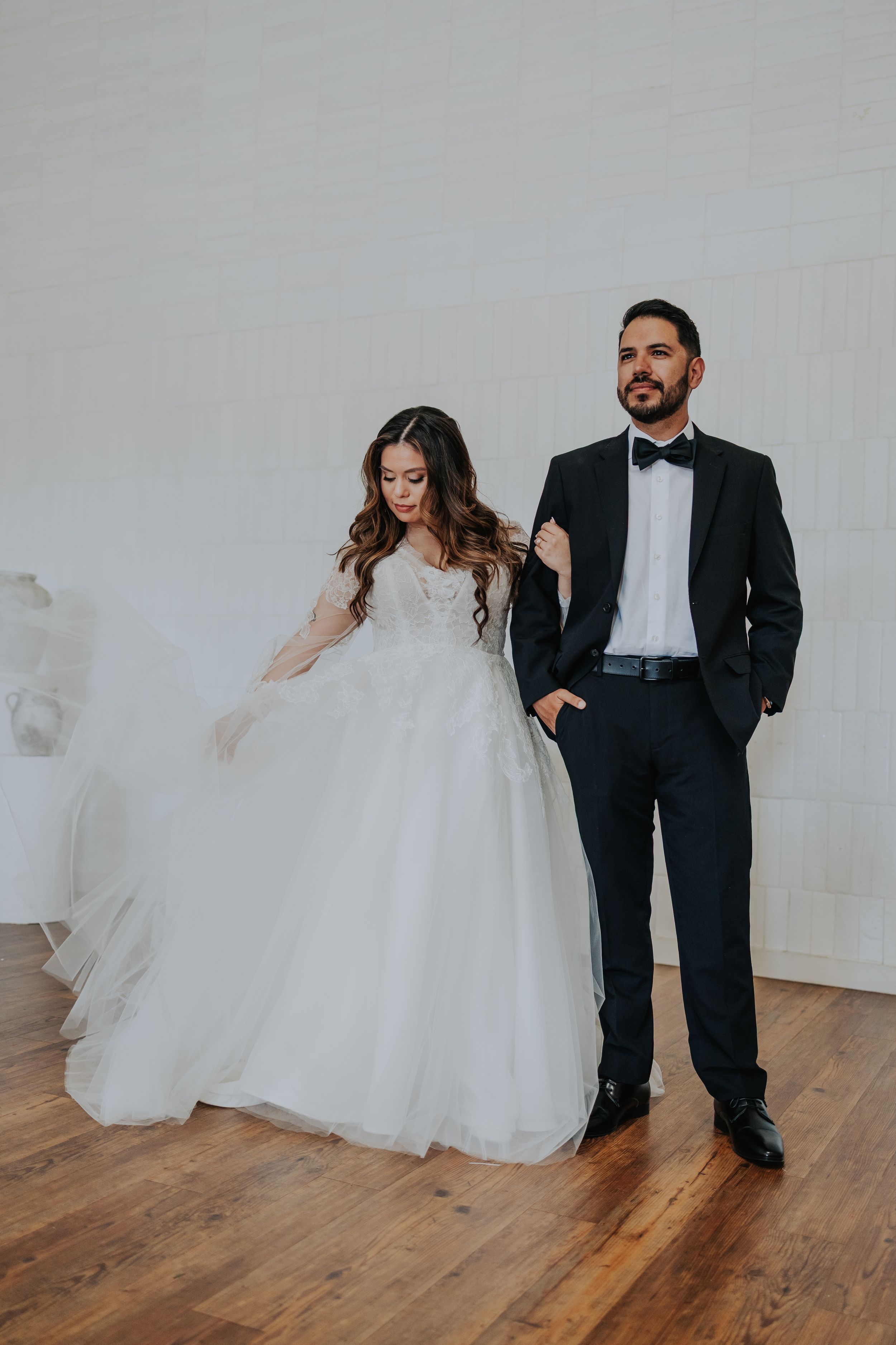 Bride in white ball gown and groom in black tuxedo posing together on hardwood floor against white wall.