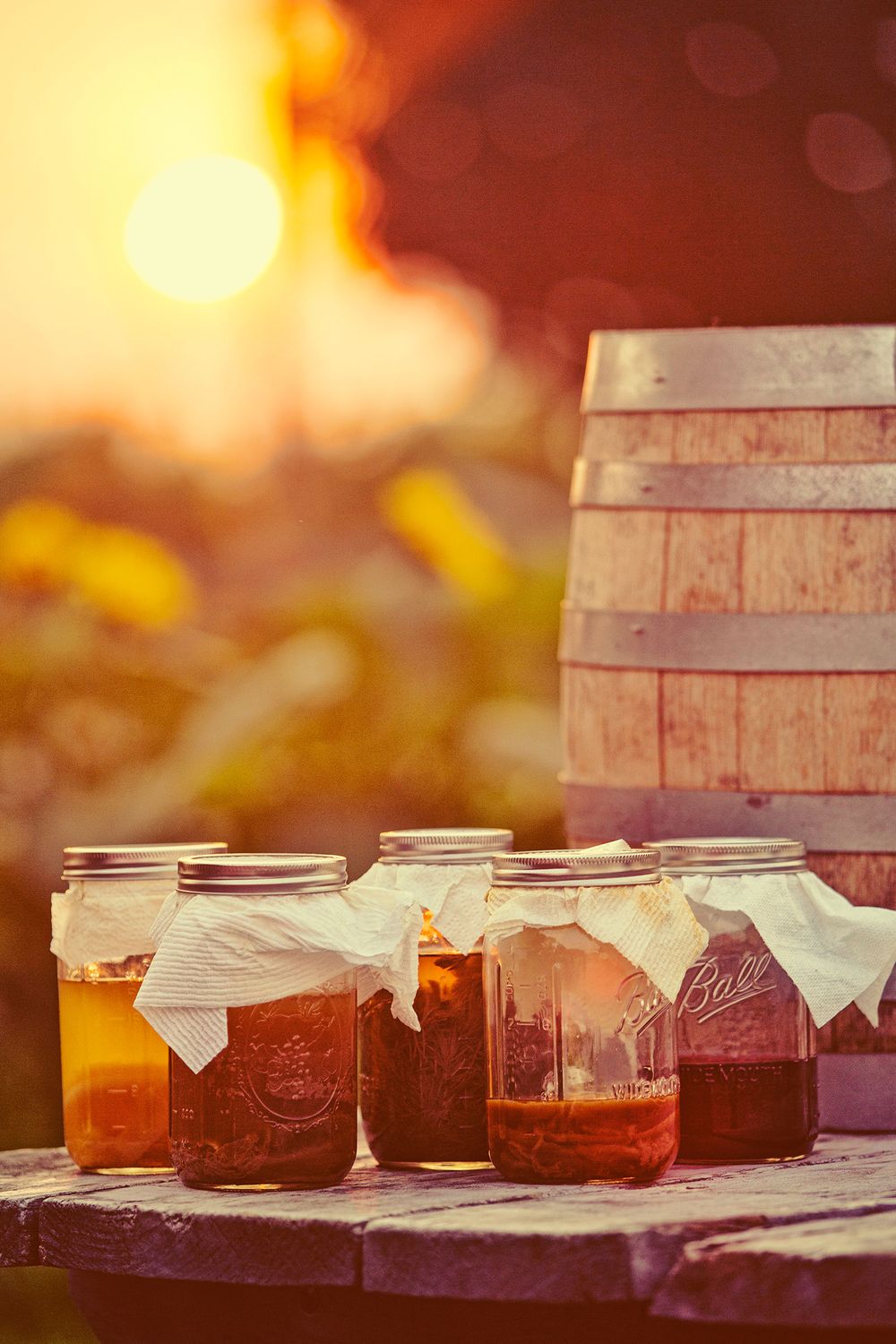 Mason jars filled with homemade kombucha on a rustic wooden table at sunset with a barrel.