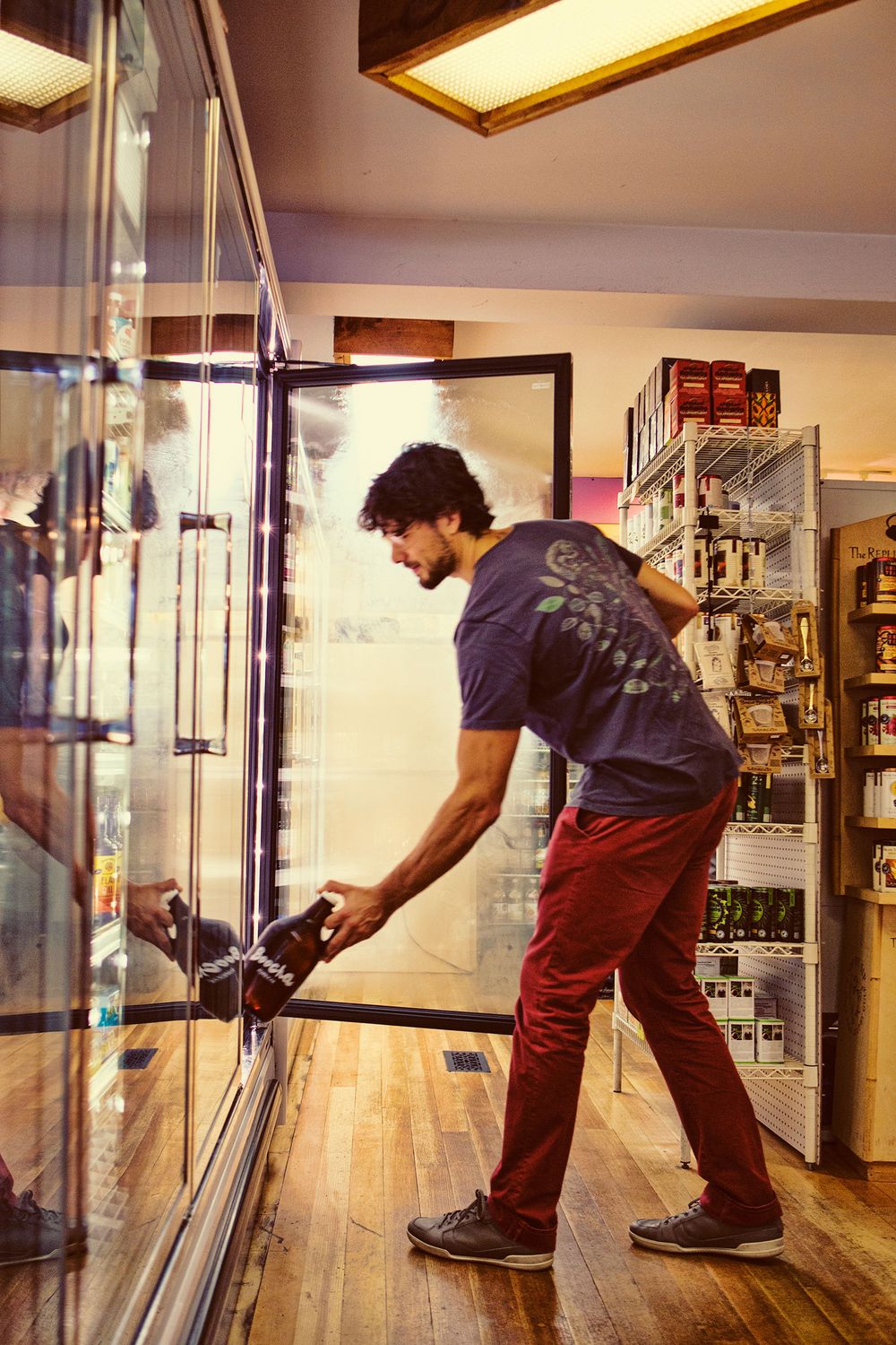 Young man in red pants selecting a kombucha bottle from a refrigerated store display case.