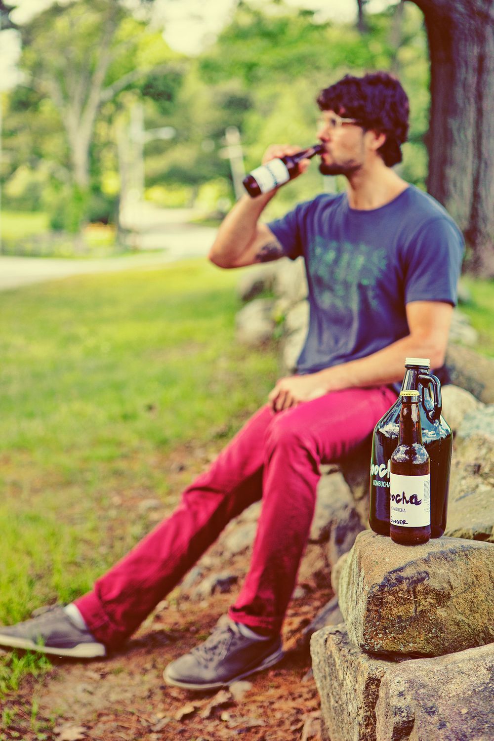 Man sitting on rocks in a park drinking Boocha kombucha with a growler bottle beside him.