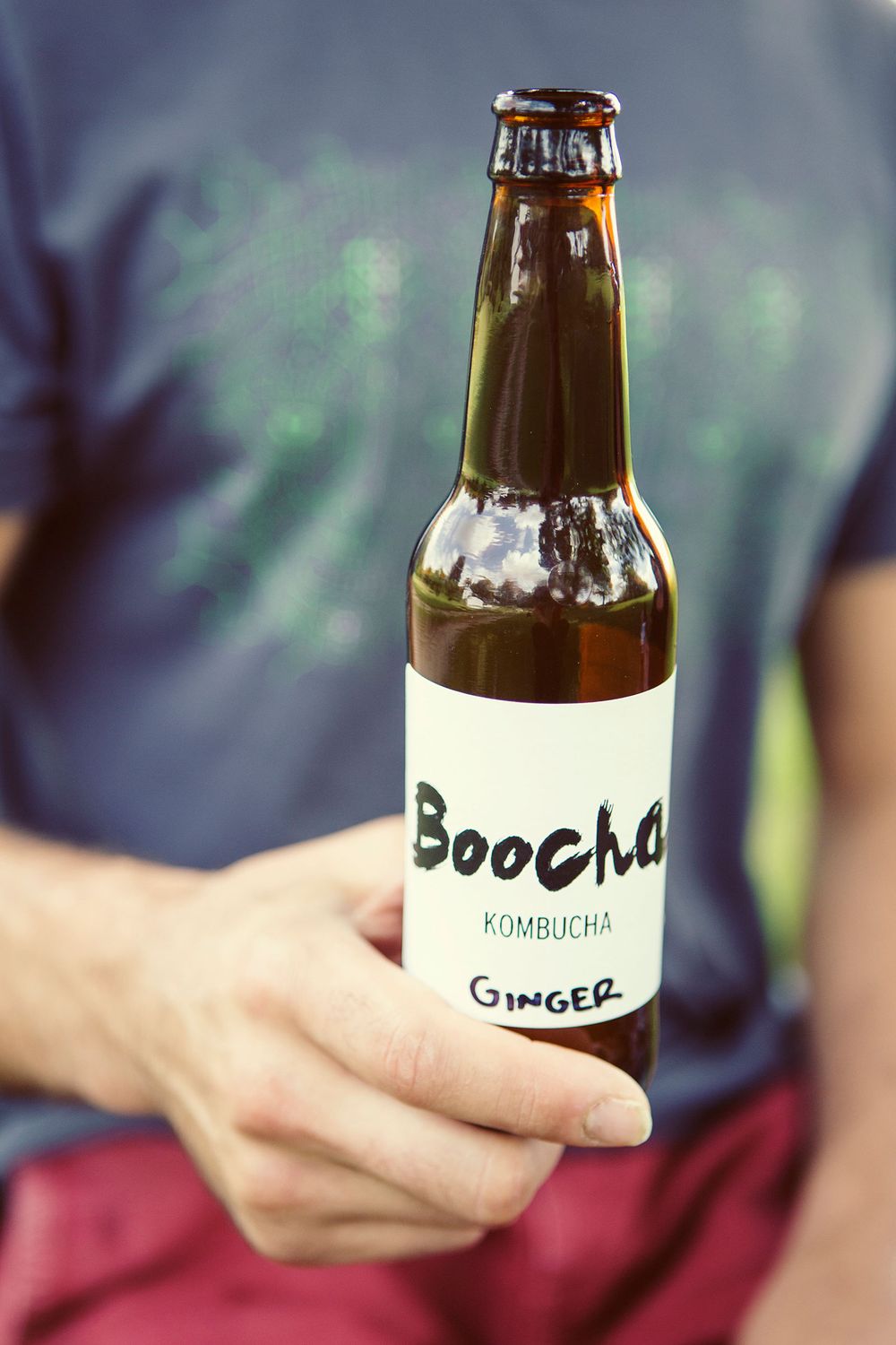 Hand holding a brown bottle of Boocha Ginger Kombucha outdoors.