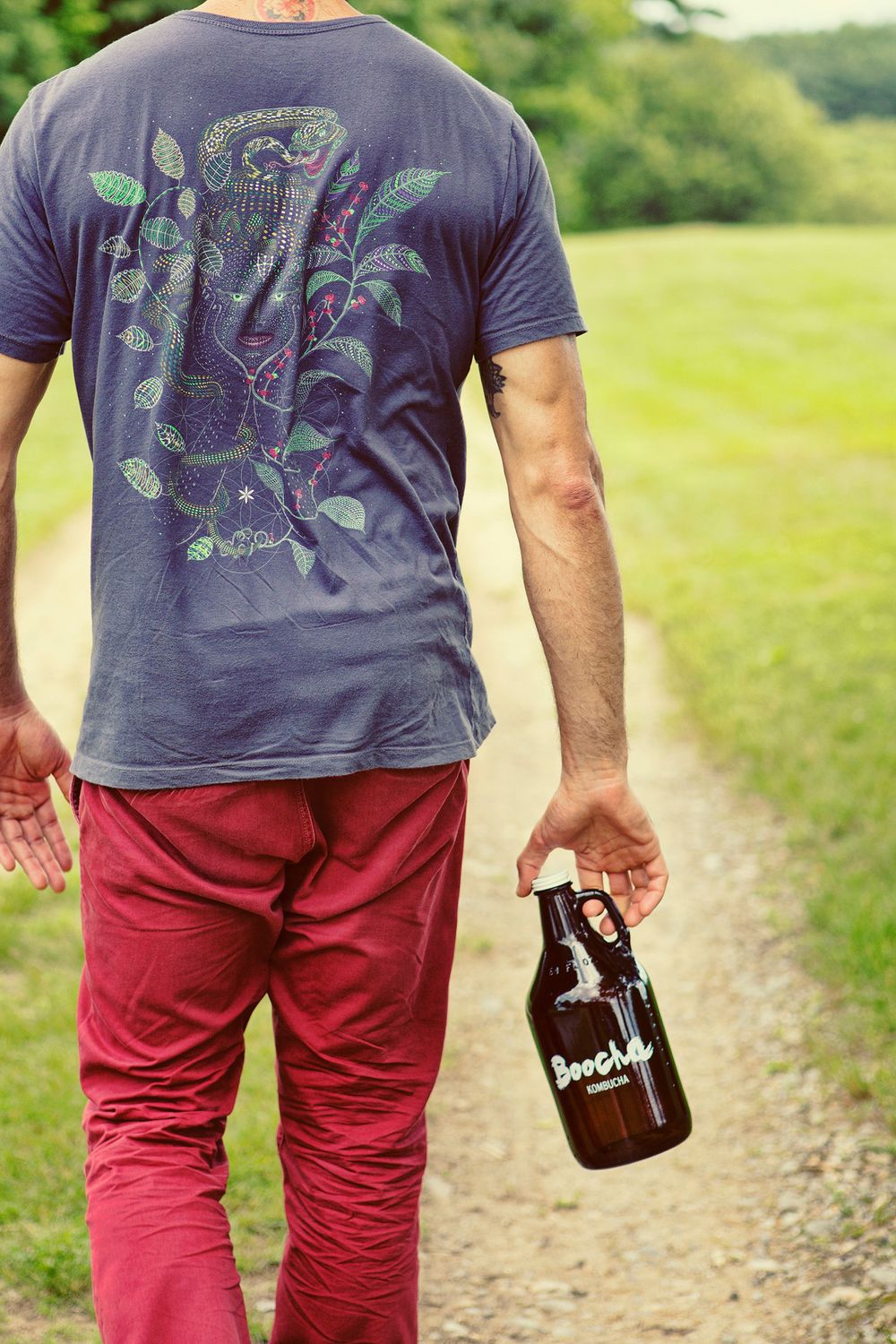 Man in graphic tee and red pants walking on a dirt path holding a Boocha growler.