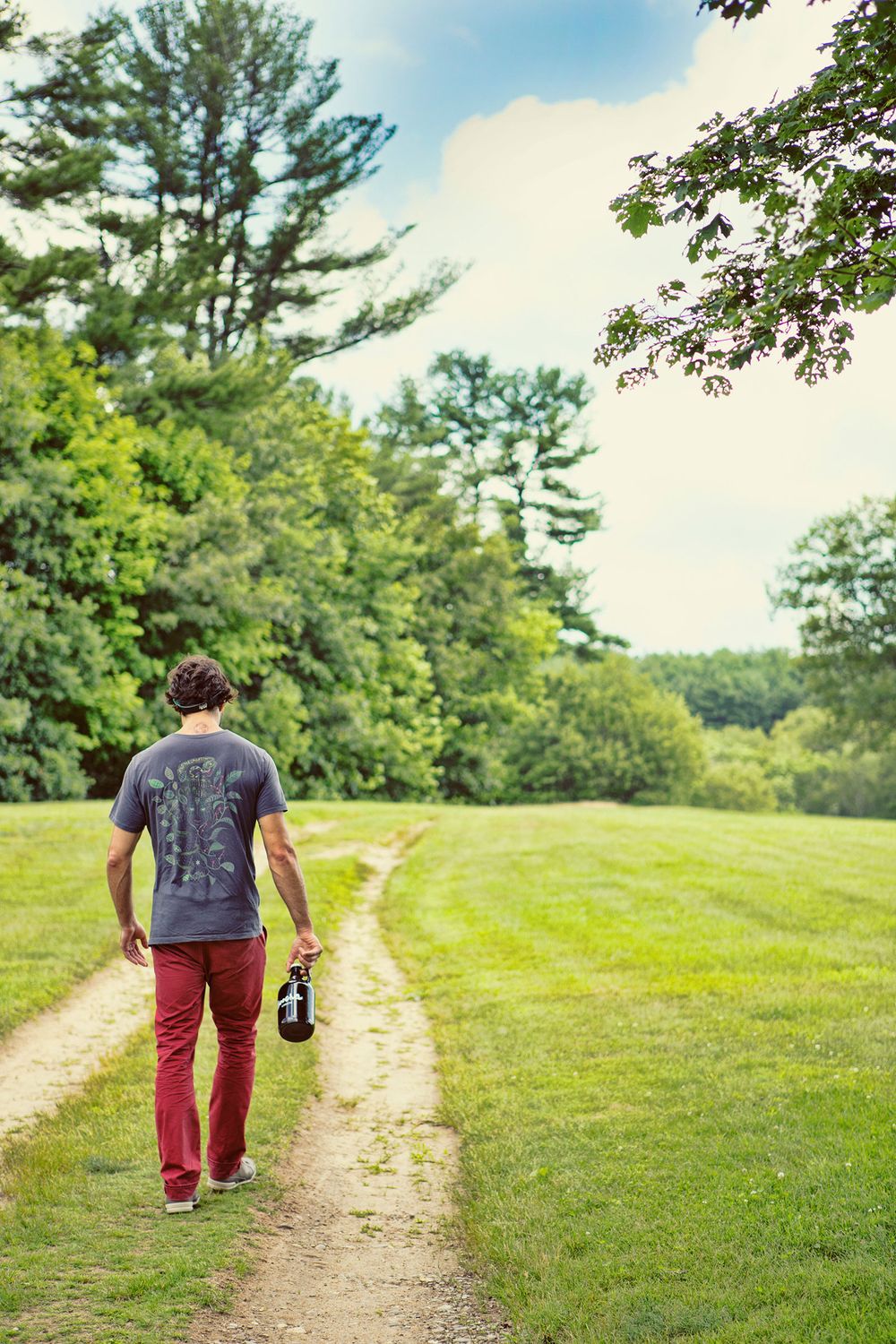 Man walking away on a scenic green park path carrying a dark Kombucha growler.