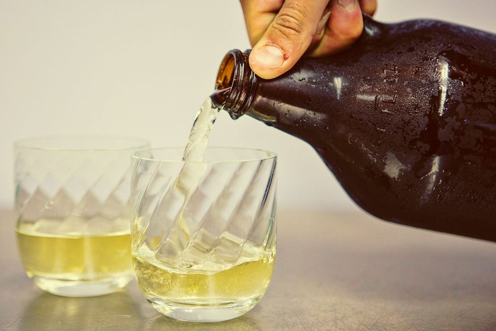 Pouring golden Kombucha from a brown bottle into two crystal glasses.
