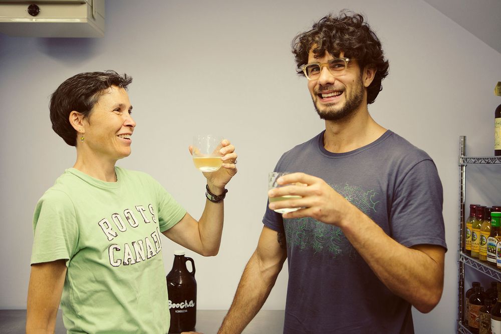 Two smiling people toasting with glasses of Boocha Kombucha indoors.