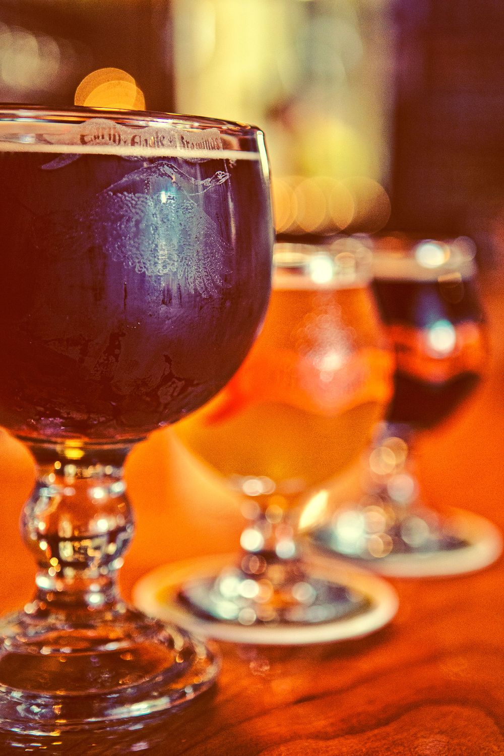 Row of craft beer goblet glasses on a wooden bar with warm bokeh lighting.