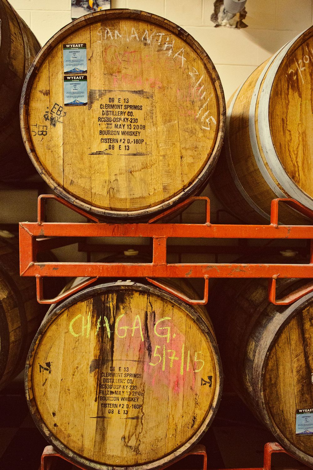 Stacked oak whiskey barrels on red metal racks in a distillery warehouse with handwritten labels.