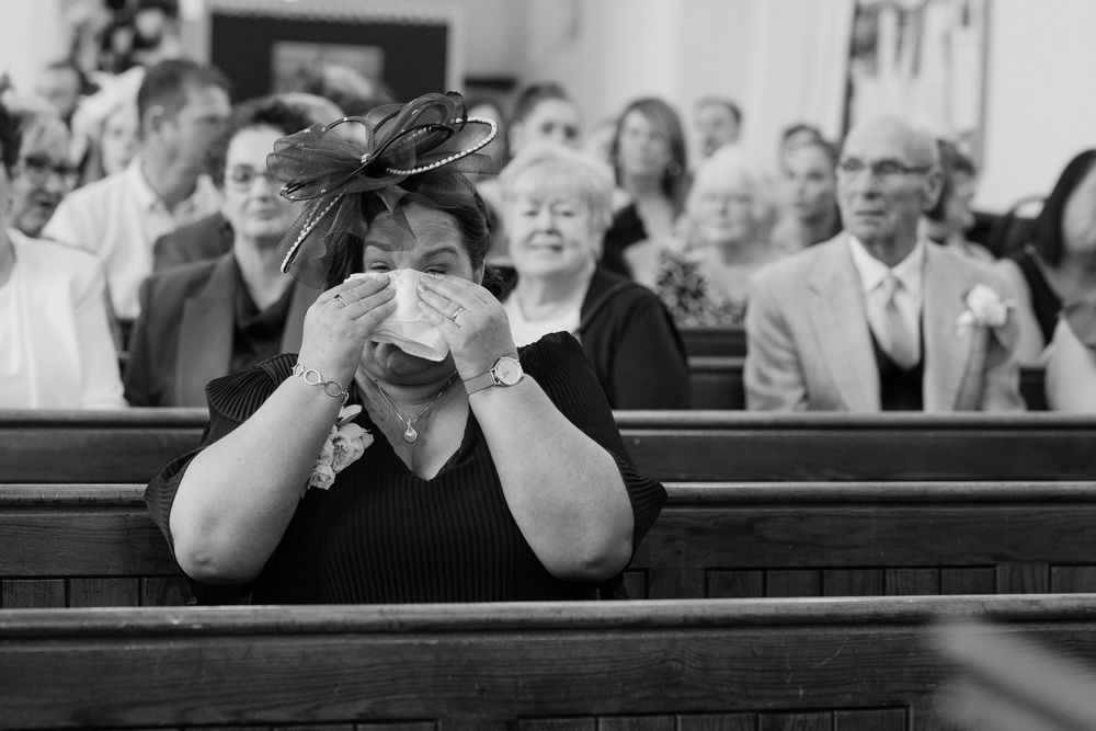 Emotional wedding guest crying into tissue in church pew during ceremony, black and white photo.