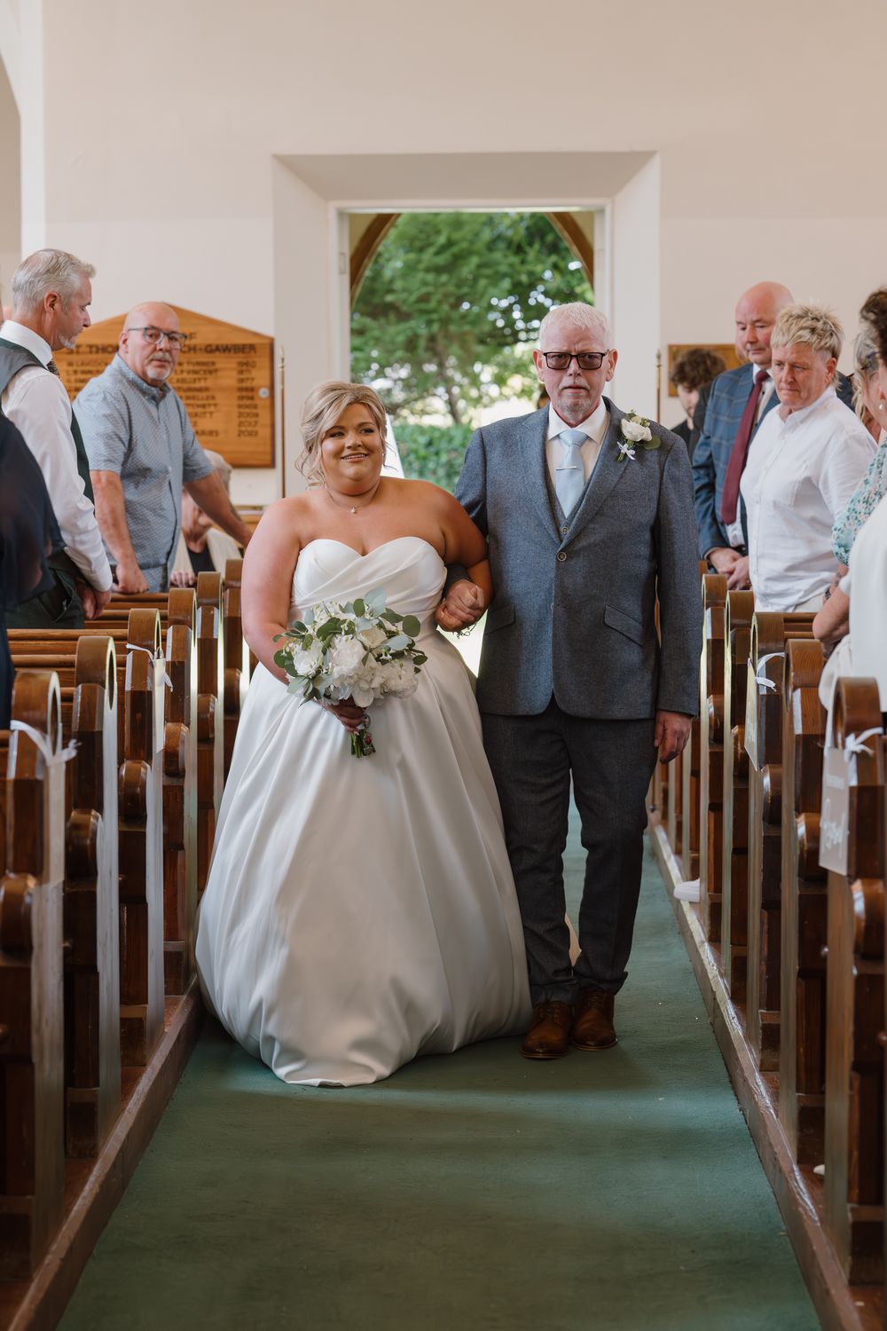 Bride in white ball gown walks down church aisle with older gentleman escort, holding bouquet.
