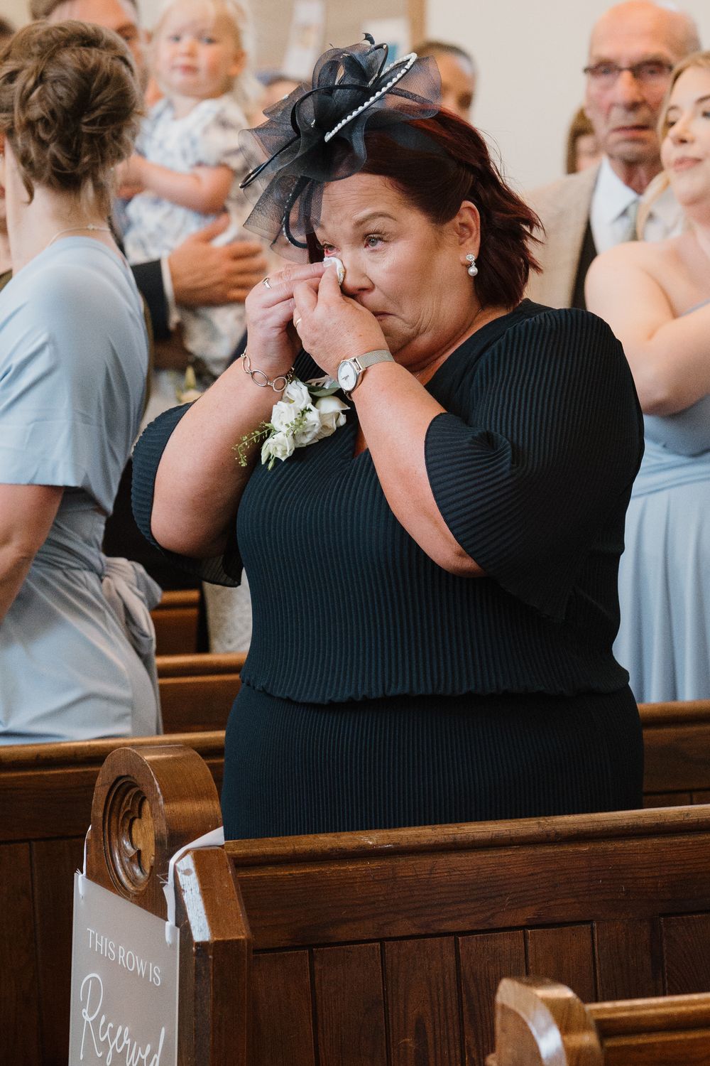 Emotional woman in black dress crying at wedding ceremony in church pew, wearing corsage and fascinator hat.
