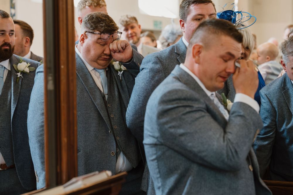 Emotional groom wiping tears at wedding ceremony with groomsmen in grey suits standing in church.