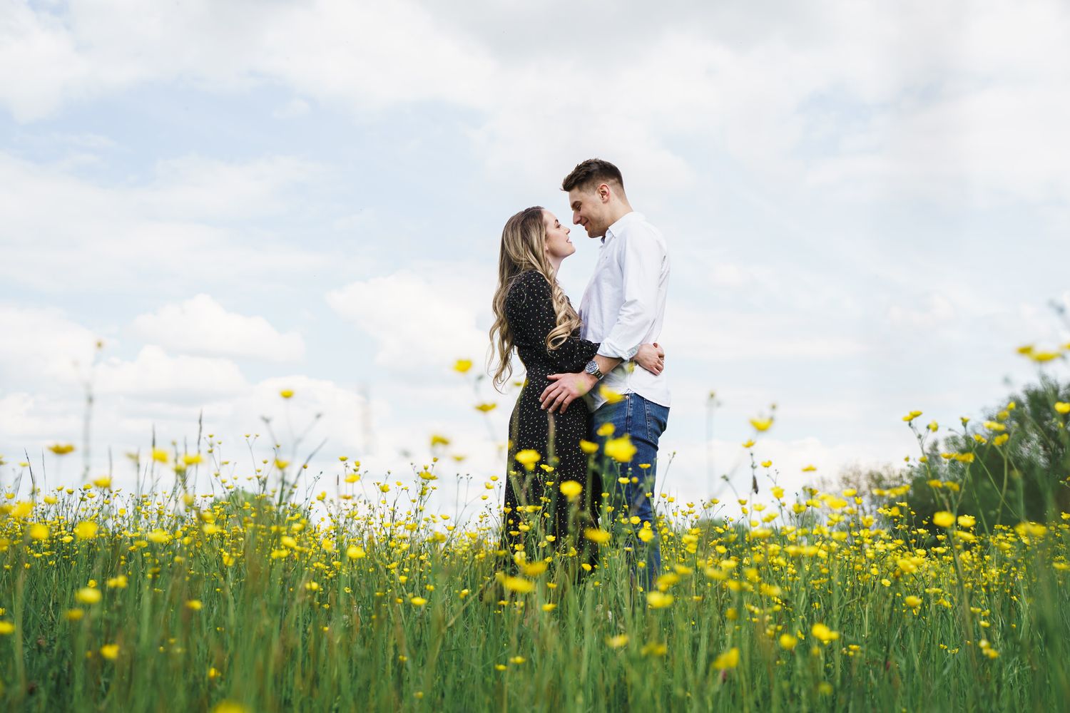 Couple embracing romantically in a field of yellow wildflowers under a partly cloudy sky.