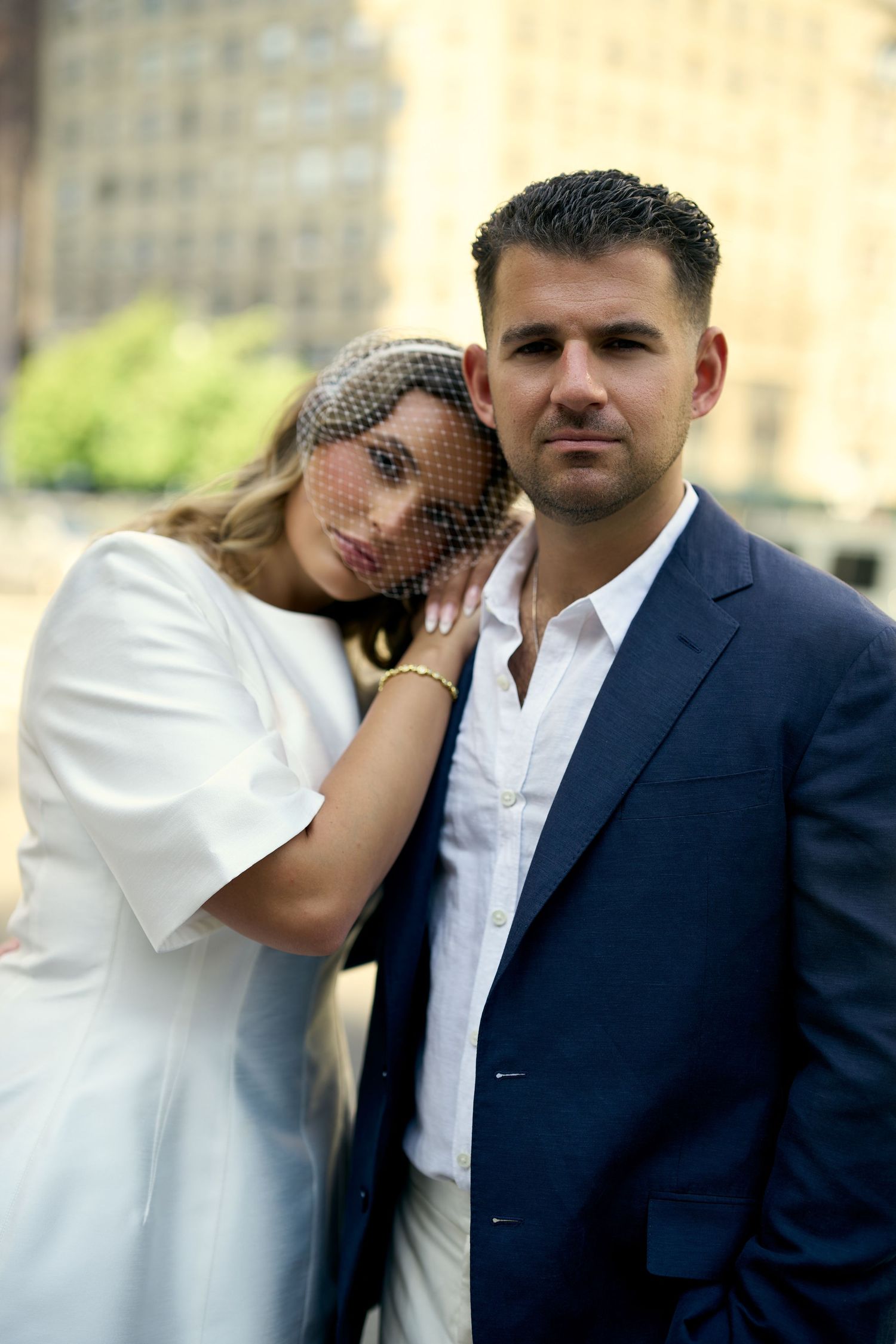 Elegant couple in wedding attire poses outdoors, bride in white dress with veil, groom in navy suit, urban backdrop.
