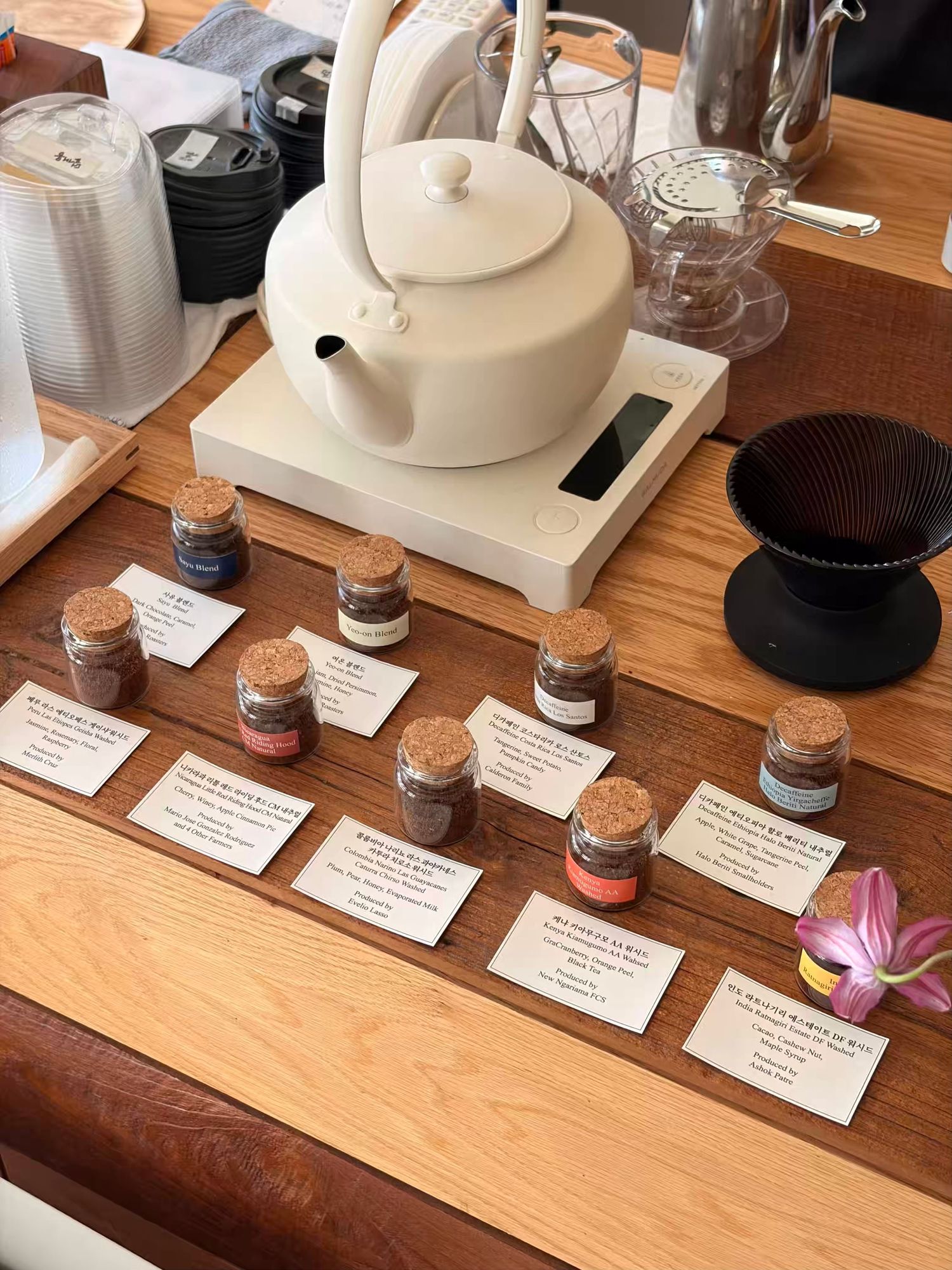 A wooden table displays small cork-topped spice jars with labels, a white ceramic teapot on a scale, and a pour-over coffee dripper.