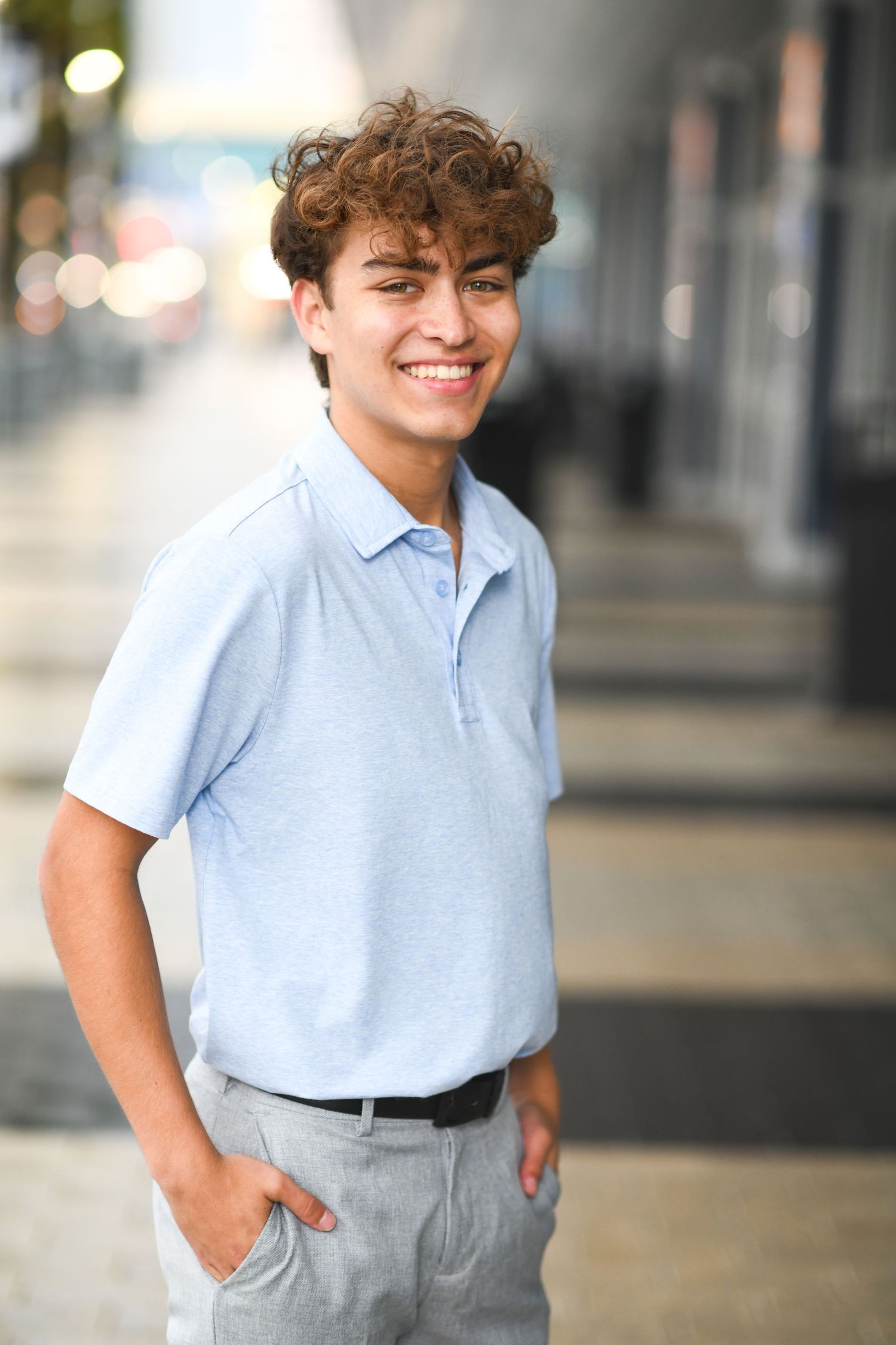 Smiling young man with curly hair wearing a light blue polo shirt and gray pants standing outdoors.