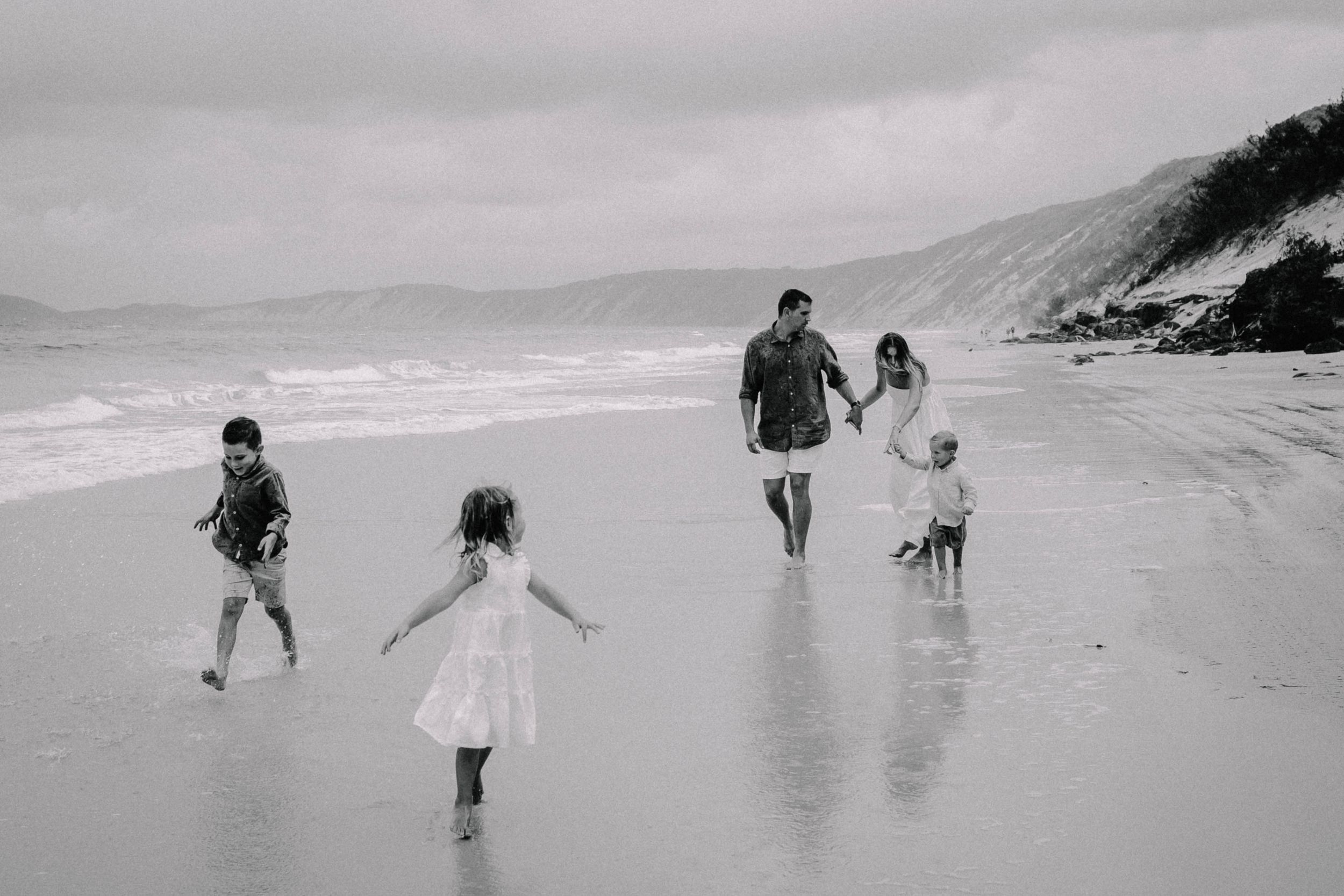 Black and white photo of a family walking on a beach with waves, two children running ahead while parents hold hands.