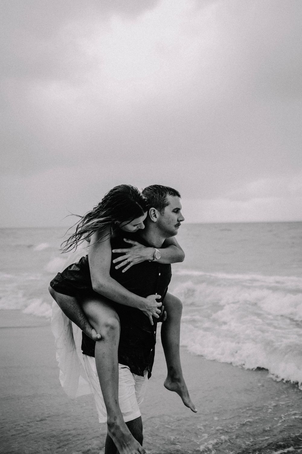 Black and white photo of a man giving a woman a piggyback ride on a beach with ocean waves in the background.