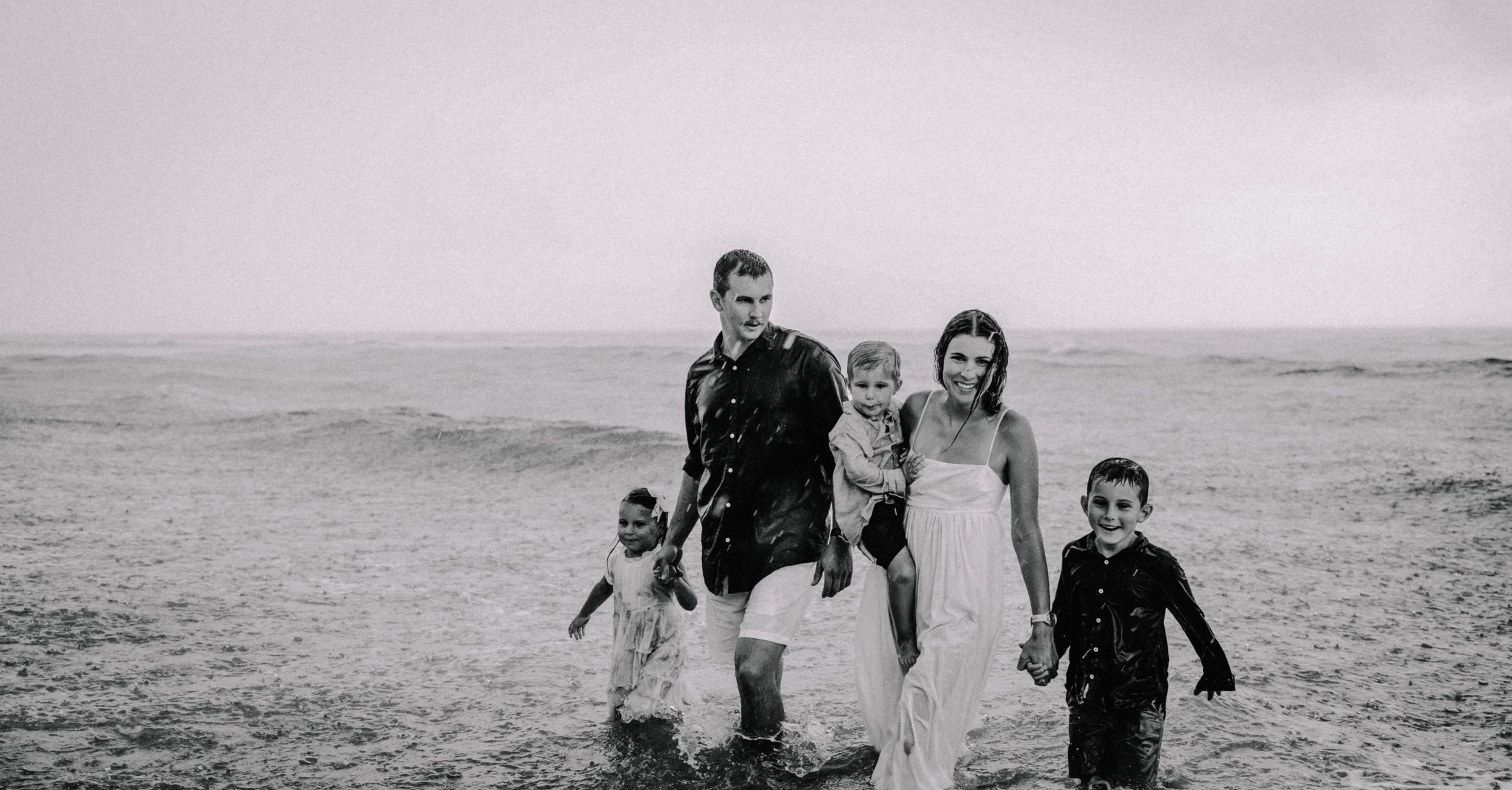 Family of five walking on a beach in black and white, parents and three children splashing in ocean waves.