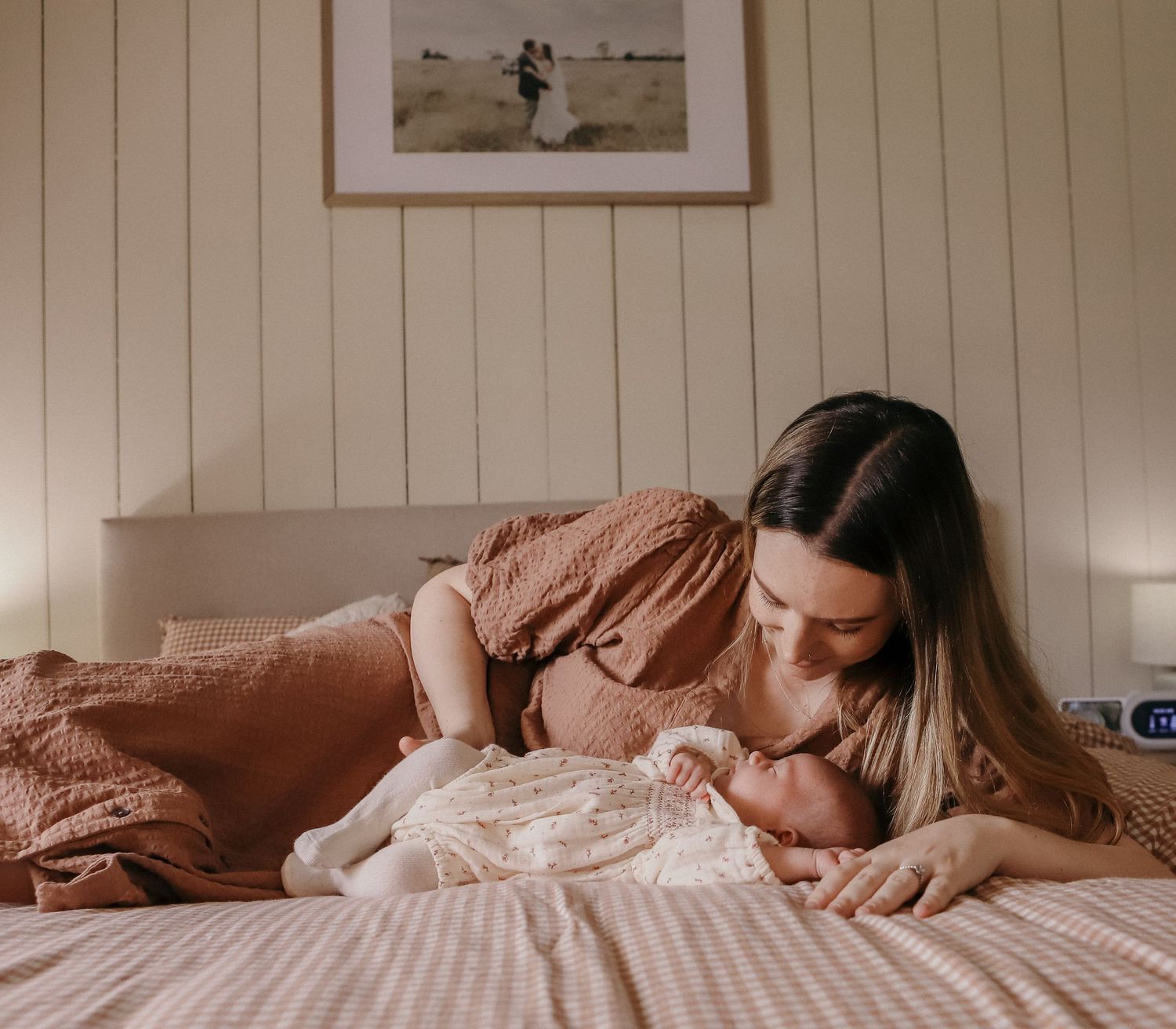 A mother tenderly gazes at her newborn baby lying on a bed, both dressed in soft earthy tones.