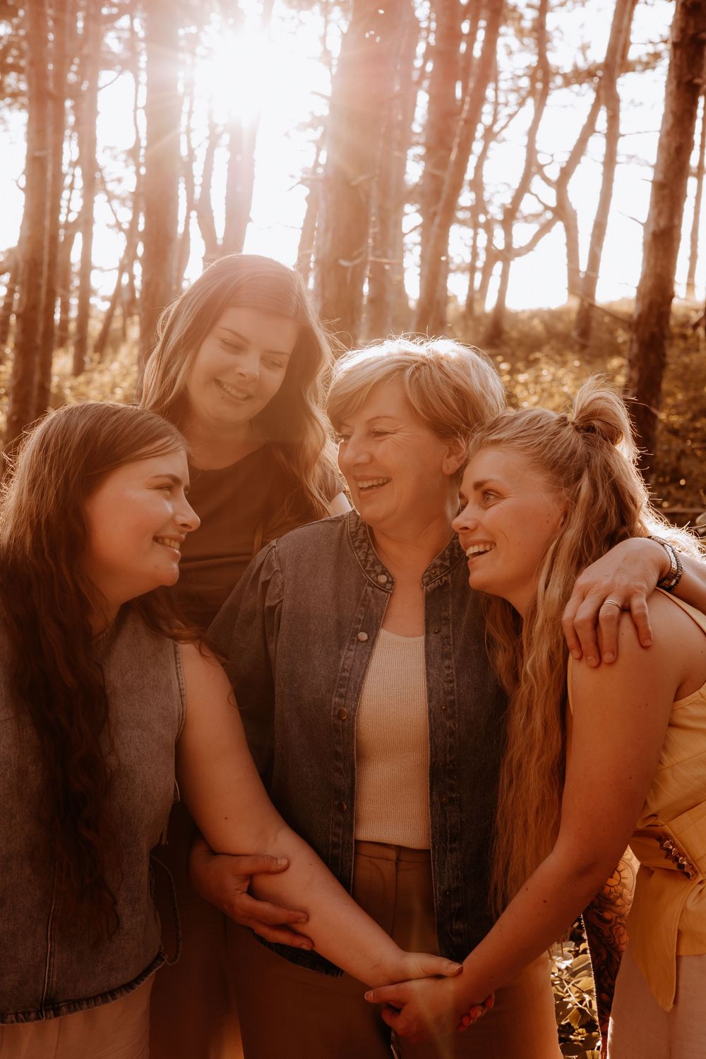 Four women smiling and embracing each other warmly in a sunlit forest at golden hour.